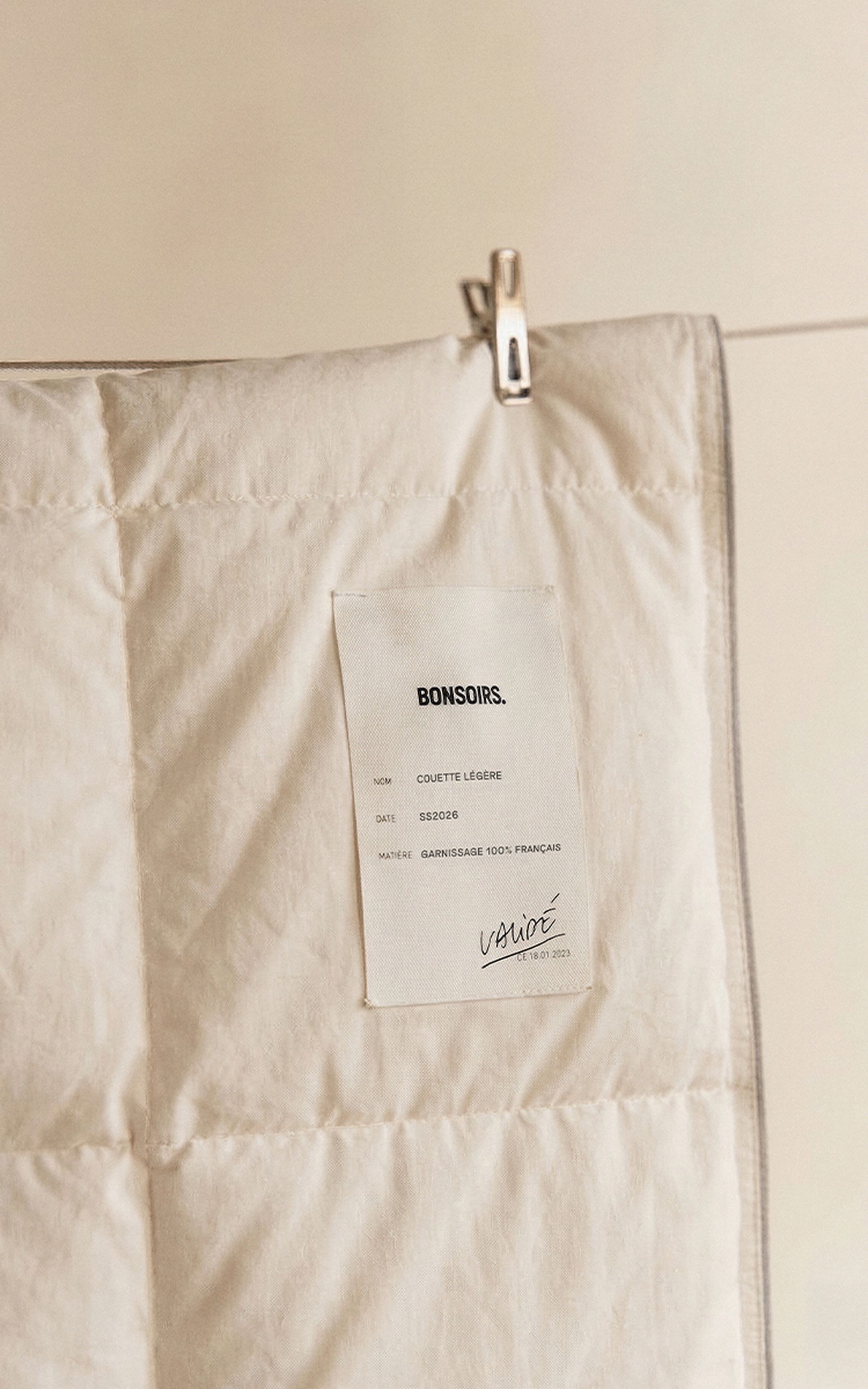 Gros plan sur une couette de couleur crème, avec une étiquette accrochée à une corde, sur laquelle figurent des informations telles que le nom, la date et la composition.