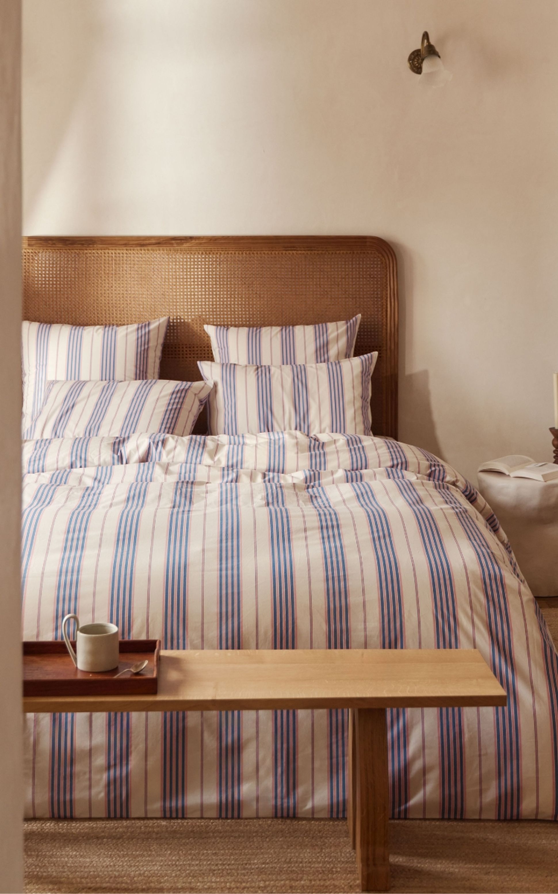 Chambre confortable avec une tête de lit en rotin et un lit soigneusement fait, recouvert d'une literie à rayures bleues. Un banc en bois et un petit plateau avec une tasse se trouvent devant.