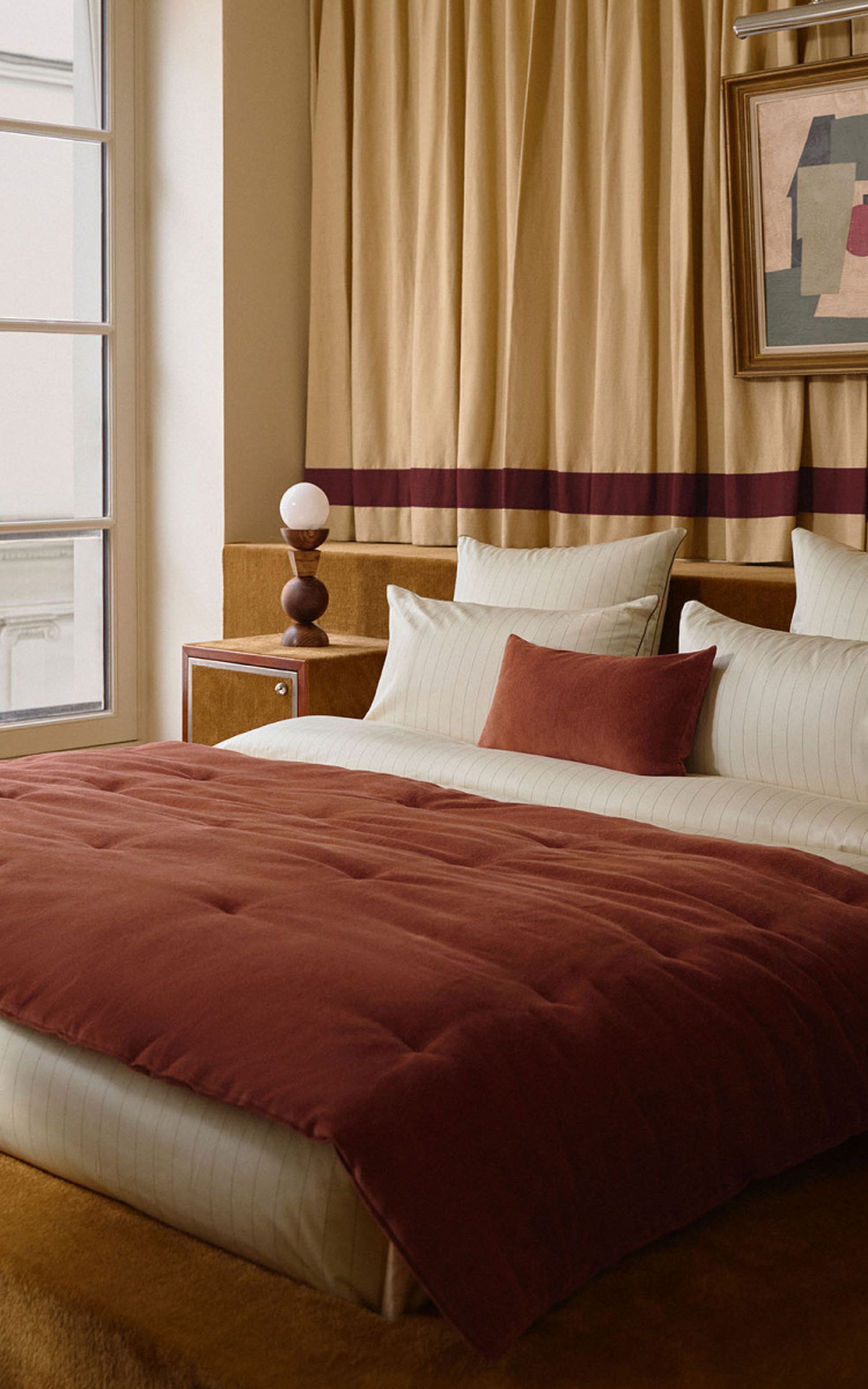 Cozy bedroom with a large bed, rust-colored bedspread, tan curtains, abstract artwork, and a lamp on a side table by the window.