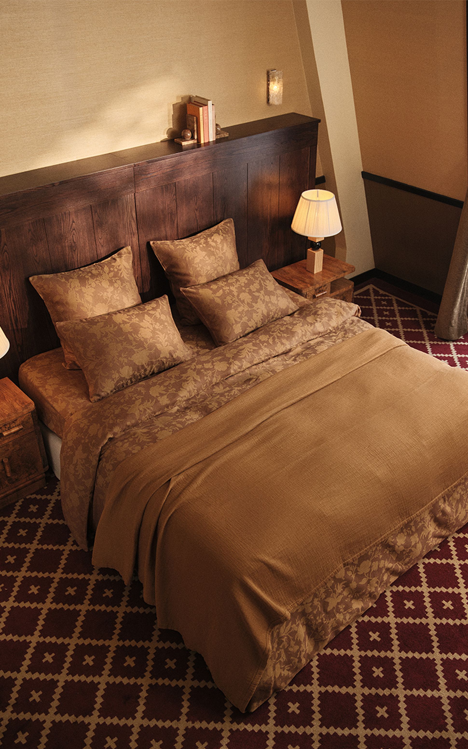 Une chambre douillette avec un grand lit recouvert d'une parure de lit à motifs marron, une tête de lit en bois, deux tables de chevet avec des lampes et des livres posés dessus.