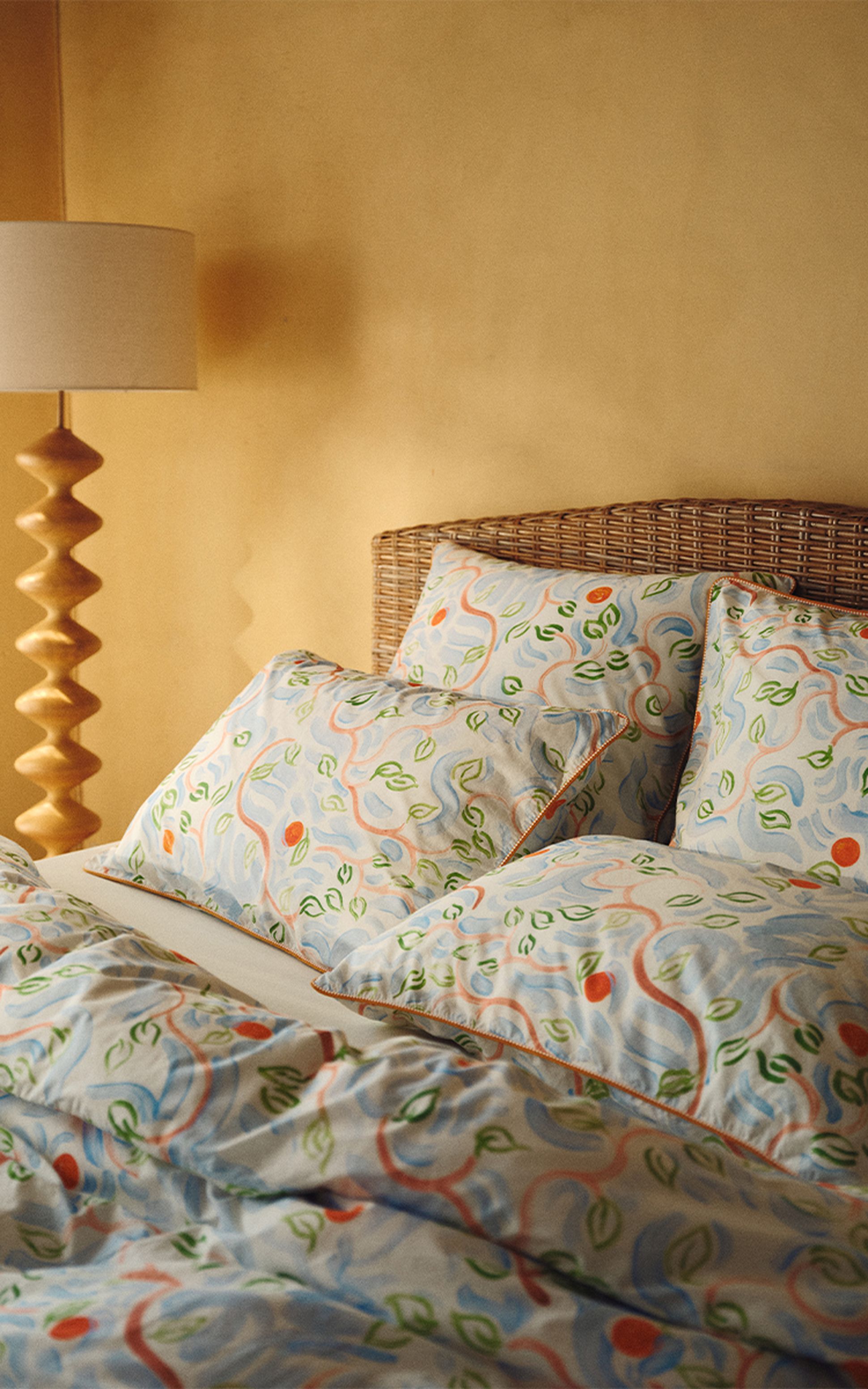 Une chambre douillette avec une tête de lit en rotin, une parure de lit aux motifs floraux colorés et une lampe élégante posée contre un mur aux tons chauds.