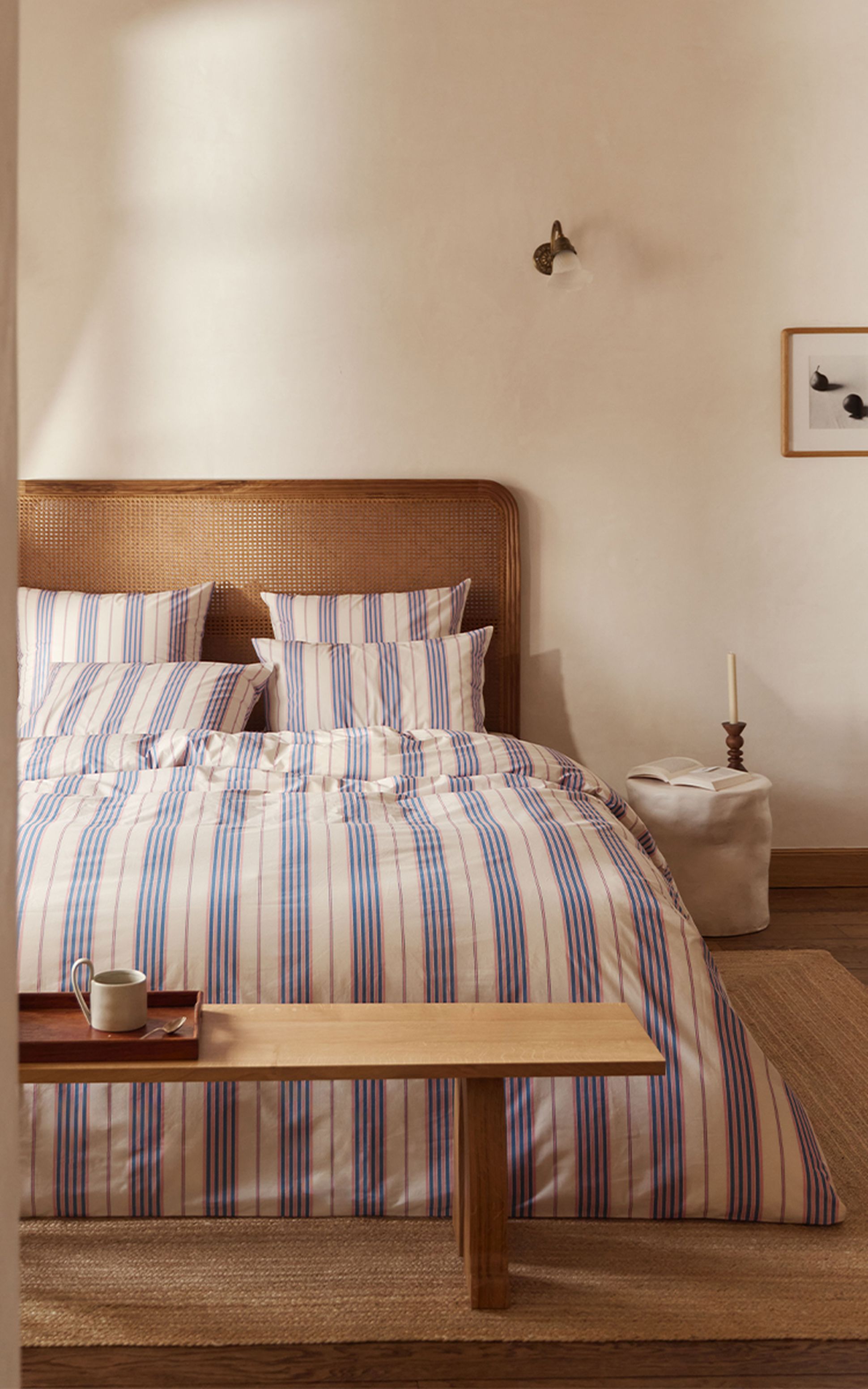 Une chambre douillette avec une parure de lit rayée, une tête de lit en bois et un banc. Une tasse et un plateau sont posés sur le banc, tandis qu'une bougie et une photo trônent sur la table d'appoint.