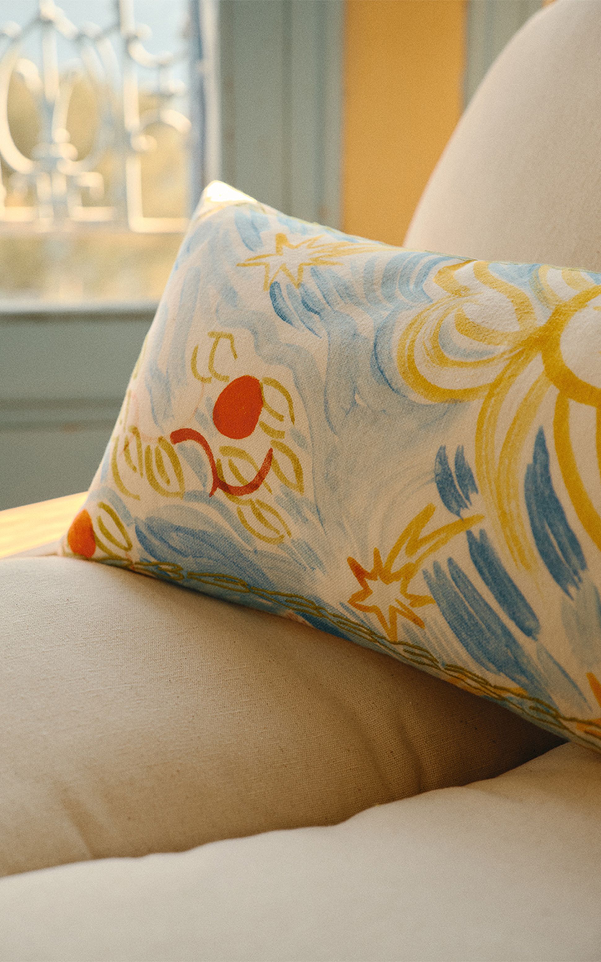 Un coussin coloré, orné de motifs floraux et abstraits, repose sur un canapé beige, baigné par la lumière chaude du soleil qui pénètre par une fenêtre voisine.