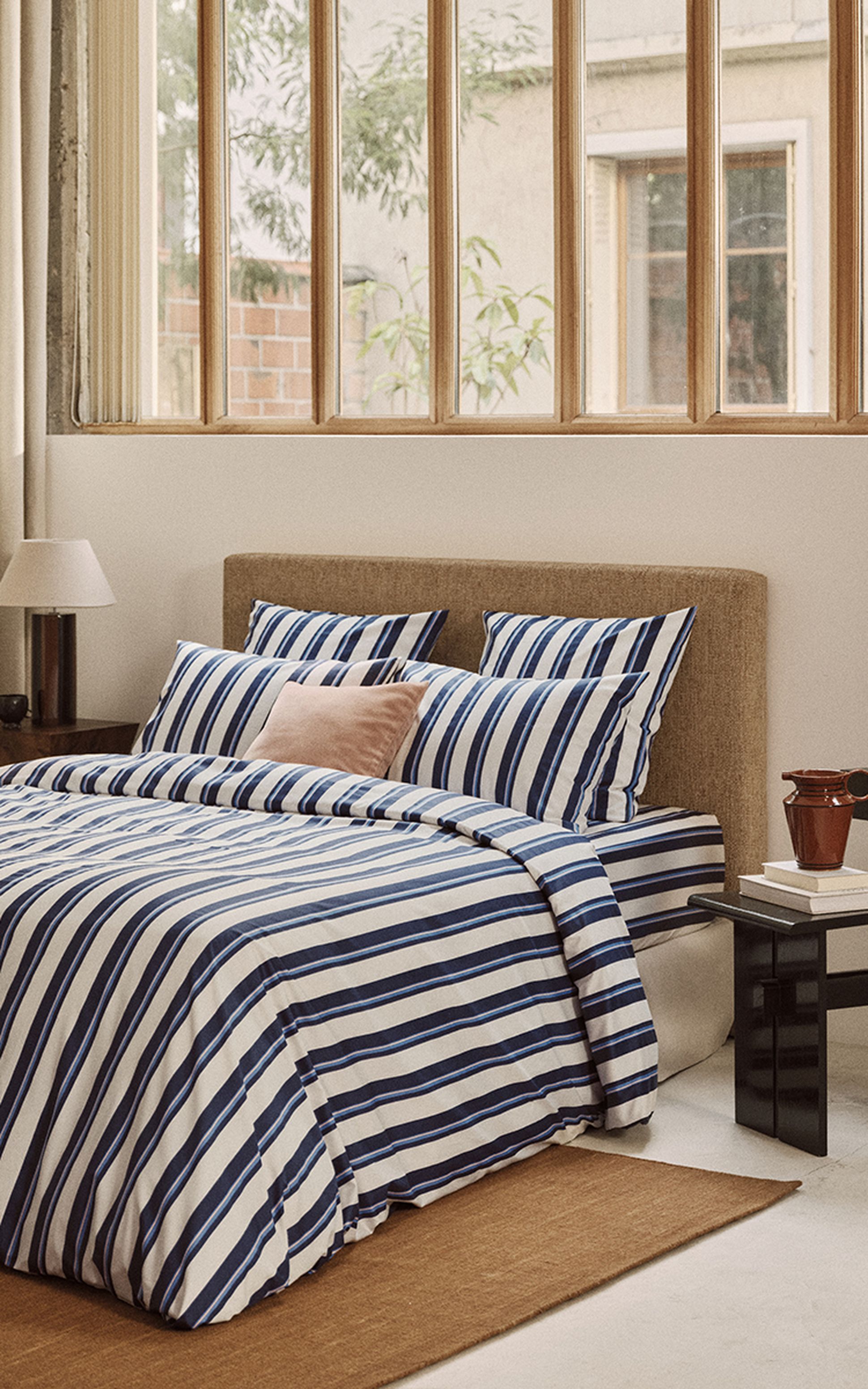 Chambre à coucher chaleureuse avec un lit habillé d'un linge de lit à rayures bleues et blanches, une tête de lit beige et une petite table de chevet sur laquelle repose une lampe, près d'une grande fenêtre.