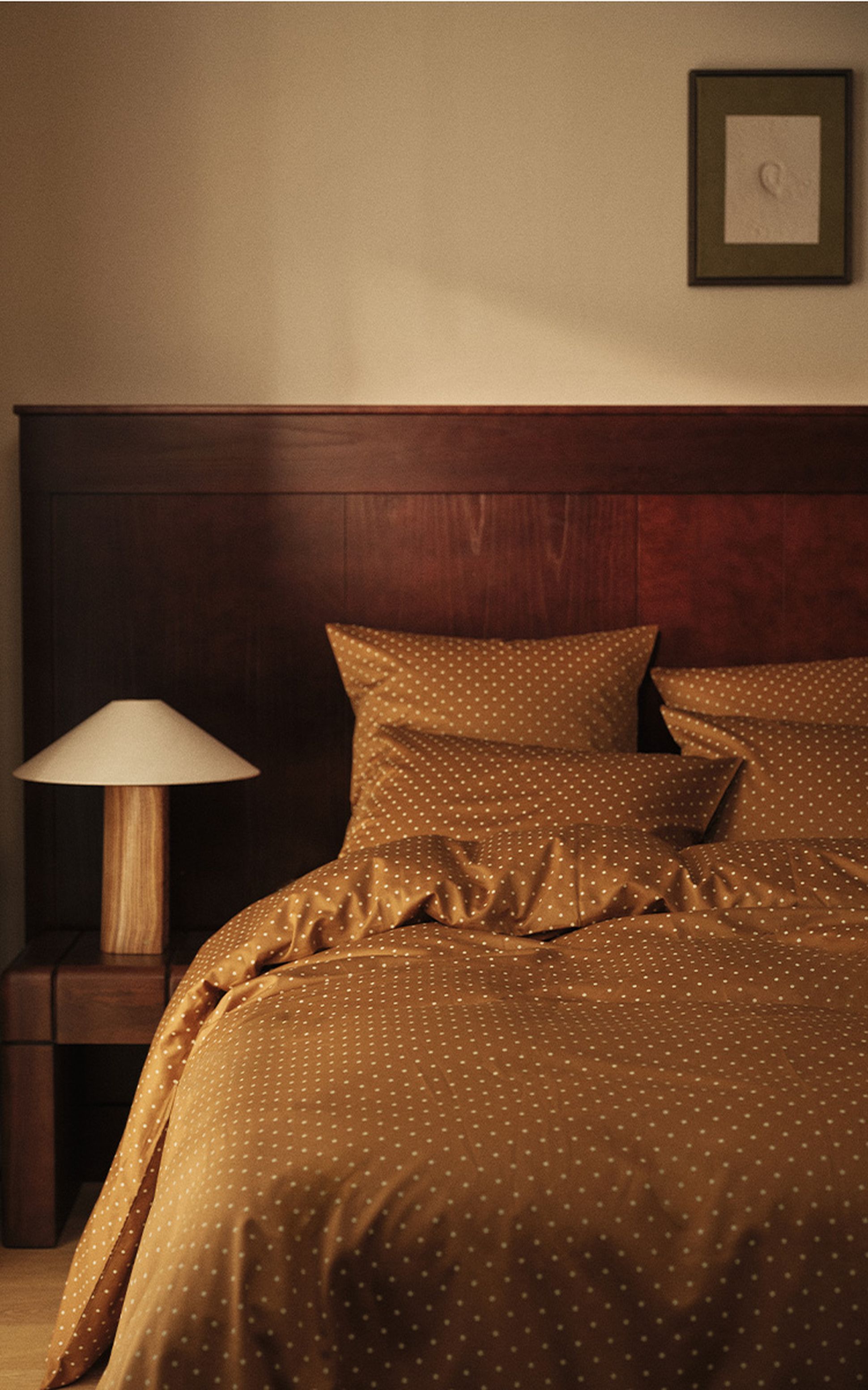 A cozy bedroom features a wooden headboard, polka-dot bedding in warm tones, a bedside lamp with a wooden base, and a framed picture on the wall.