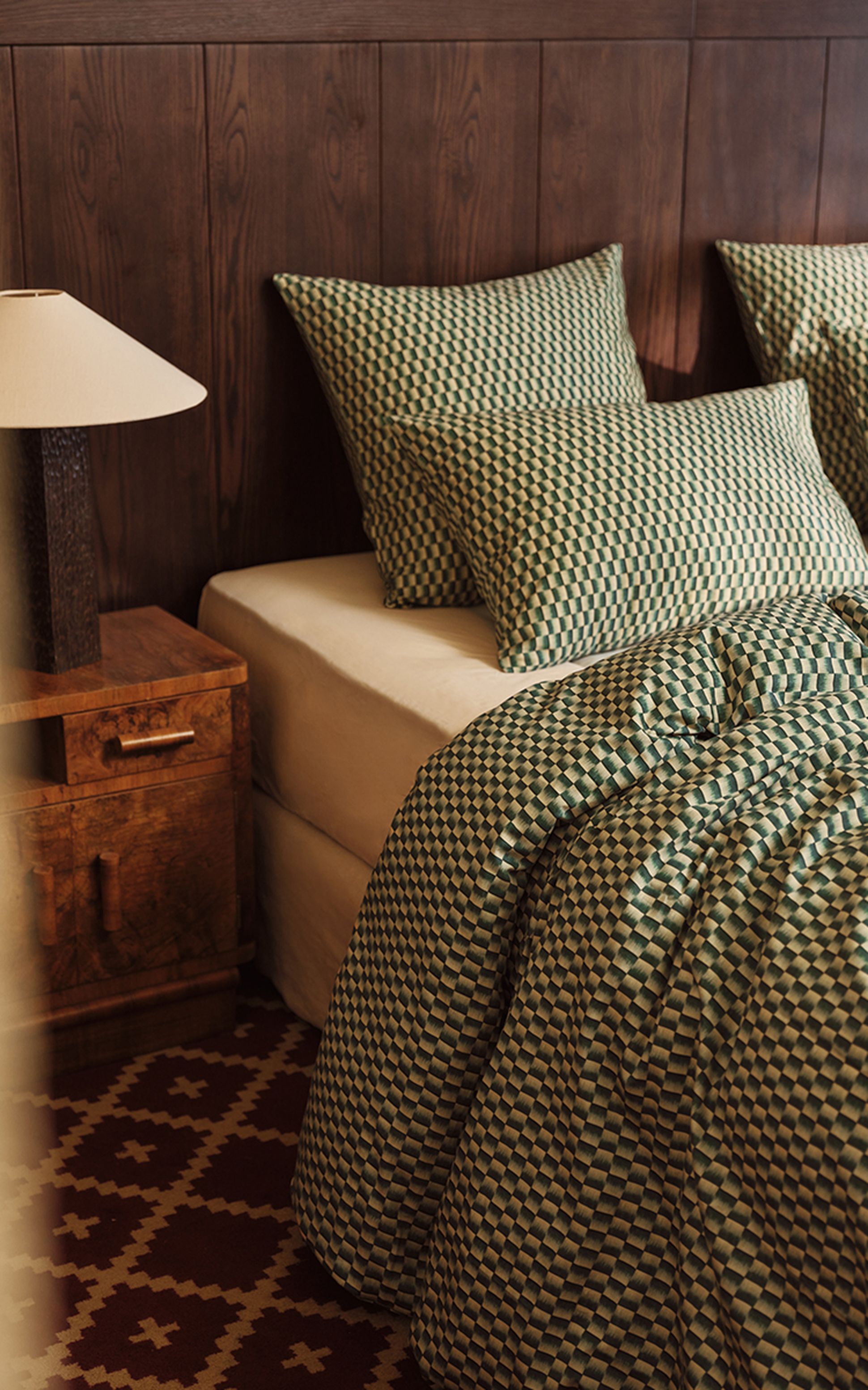 Cette chambre confortable comprend un lit avec une parure de lit à motifs verts et blancs, des oreillers assortis, une tête de lit en bois, une table de chevet et une lampe.