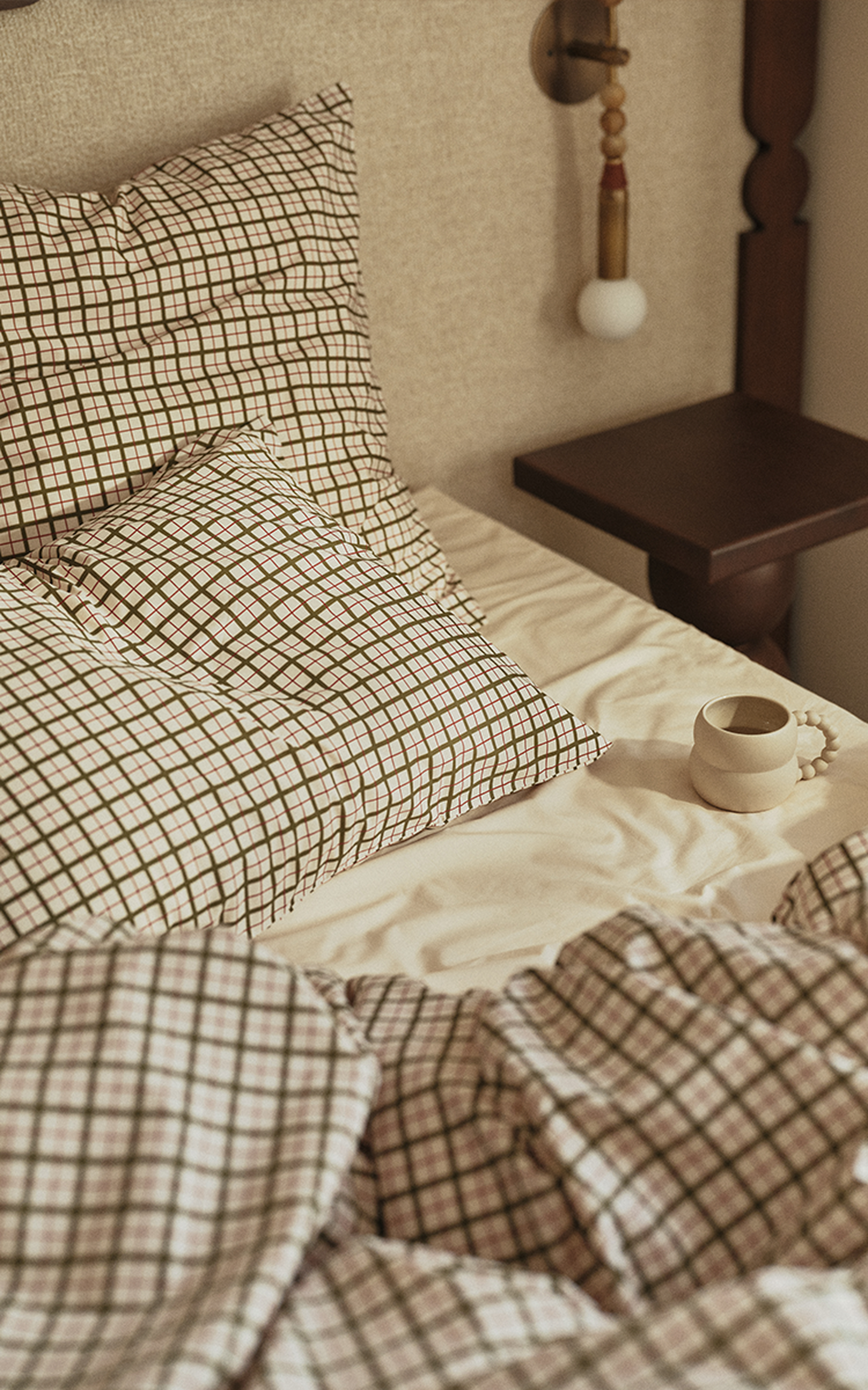 Un lit douillet avec des oreillers et une couette à carreaux, une petite table de chevet en bois et, posée à côté, une tasse blanche à l'anse ornée de perles.