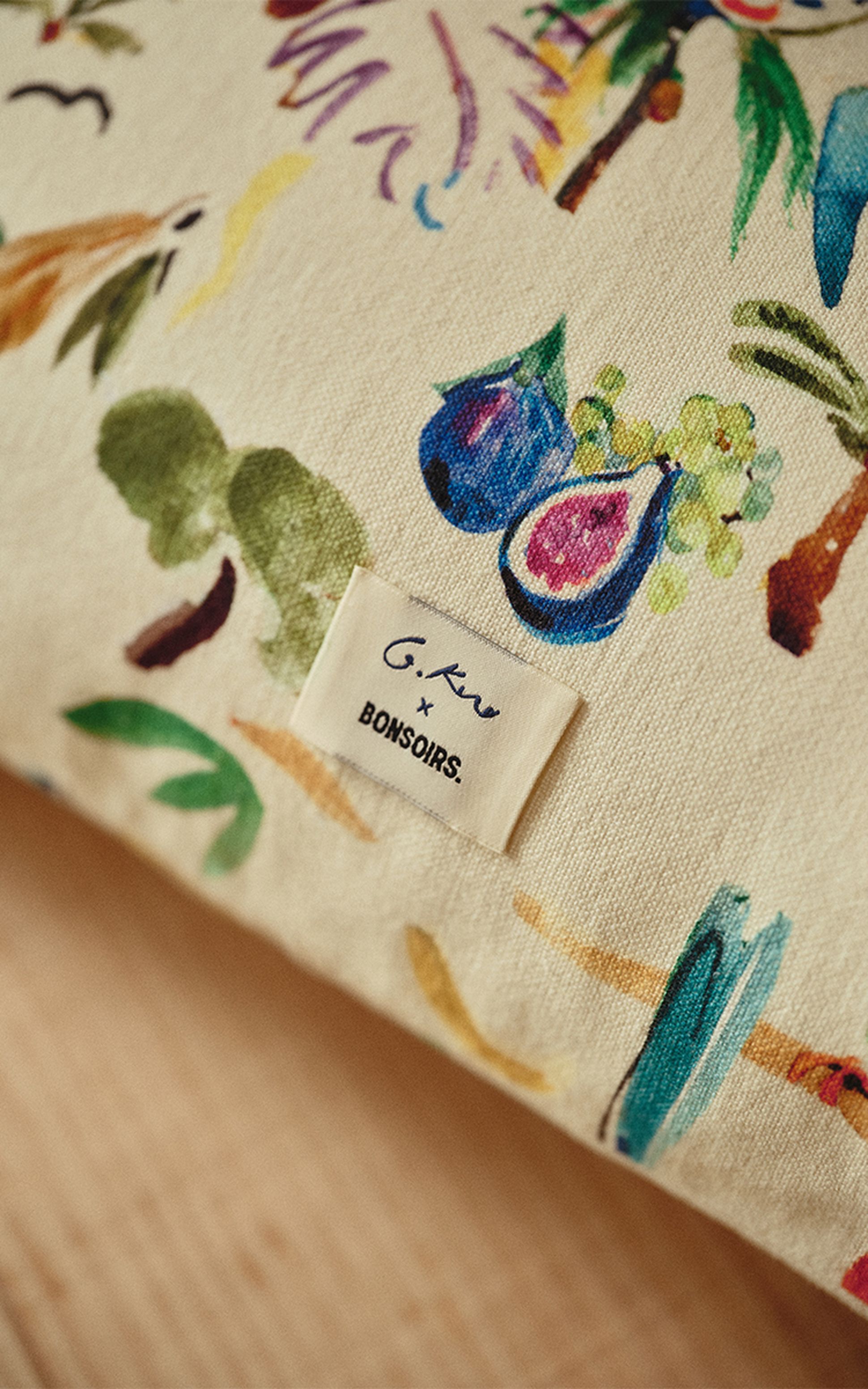 Gros plan sur un coussin coloré au motif tropical, orné d'une étiquette en tissu portant l'inscription « G.Kero x Bonsoirs », sur un fond en bois clair.