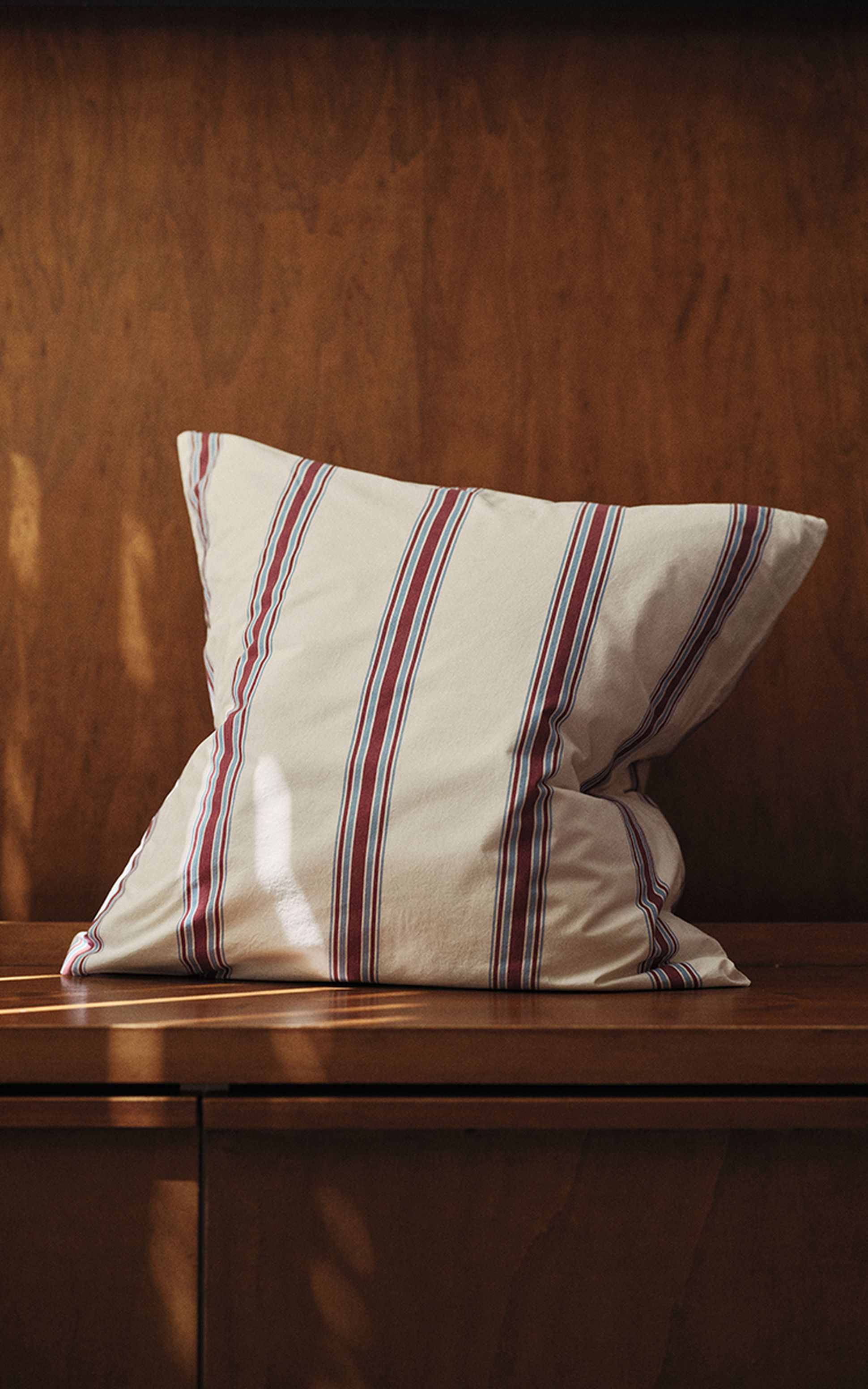 Un coussin rayé de rouge et de bleu posé sur un siège en bois, sur lequel la lumière du soleil projette des ombres douces.