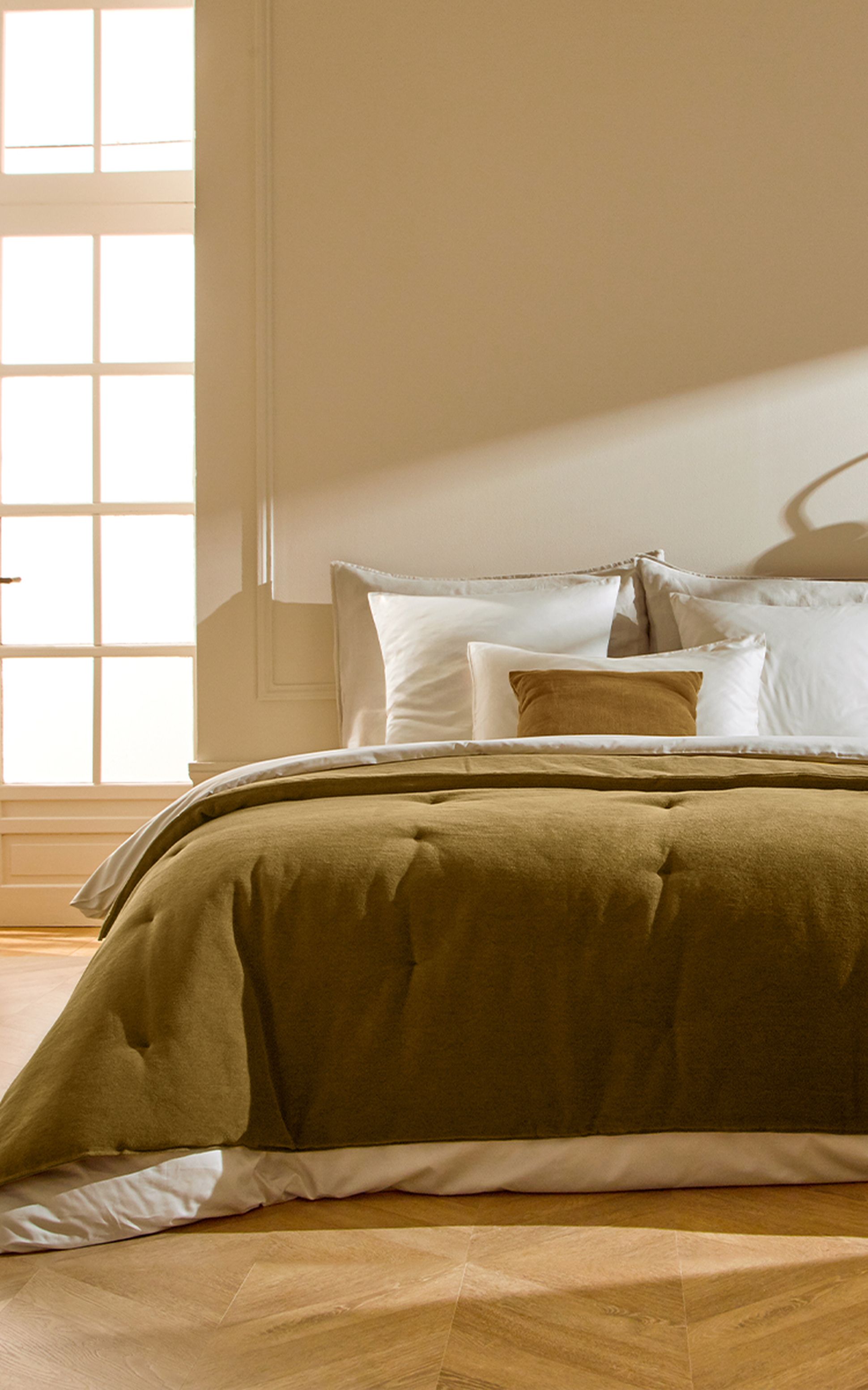 Un lit soigneusement fait, avec du linge de lit et oreillers écrus et un couvre-lit et un coussin en velours bronze, adossé à un mur clair baigné par la douce lumière du soleil qui pénètre par une grande fenêtre.