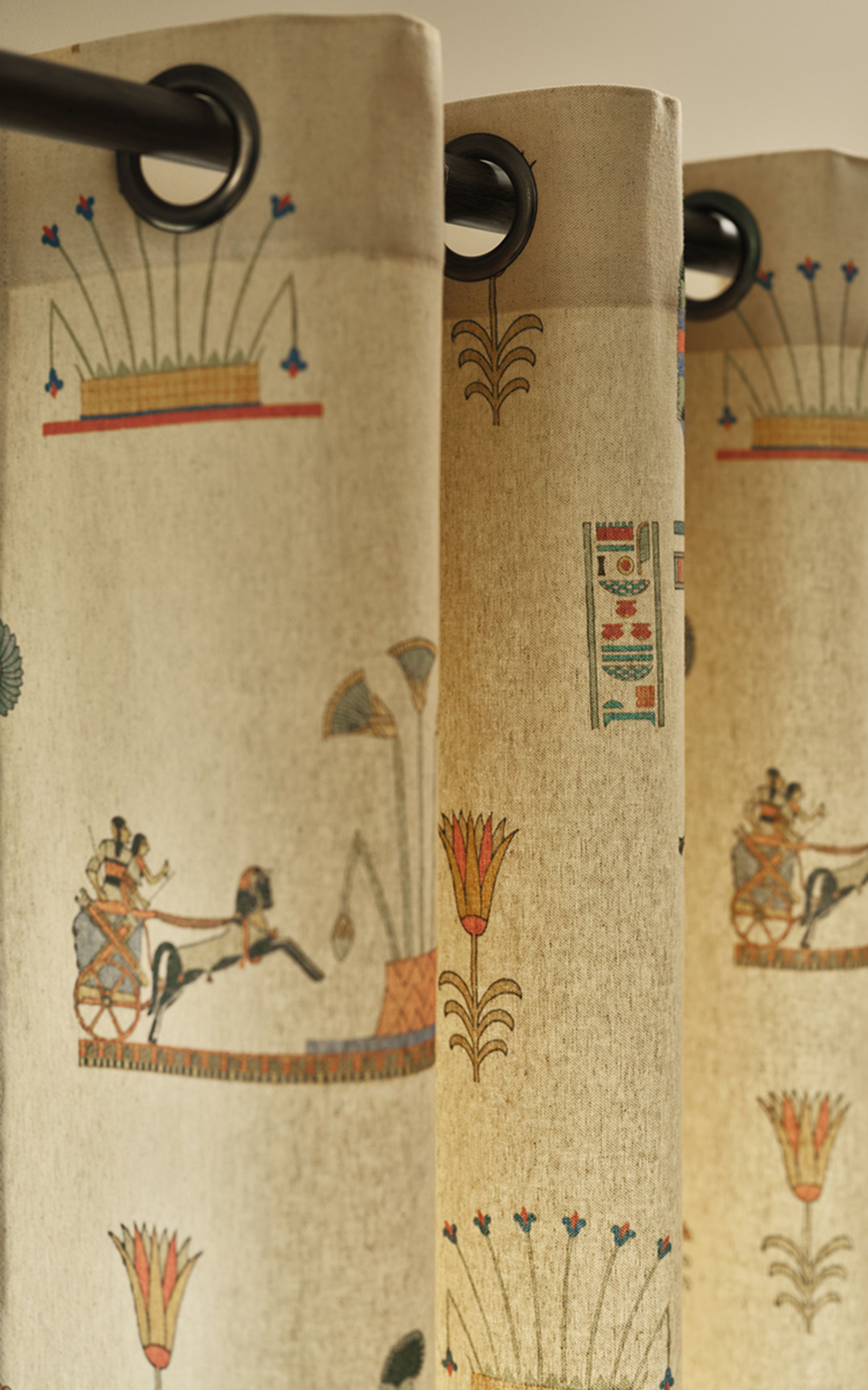Close-up of beige curtains with ancient Egyptian motifs, featuring chariots and floral designs, hung on a dark curtain rod.
