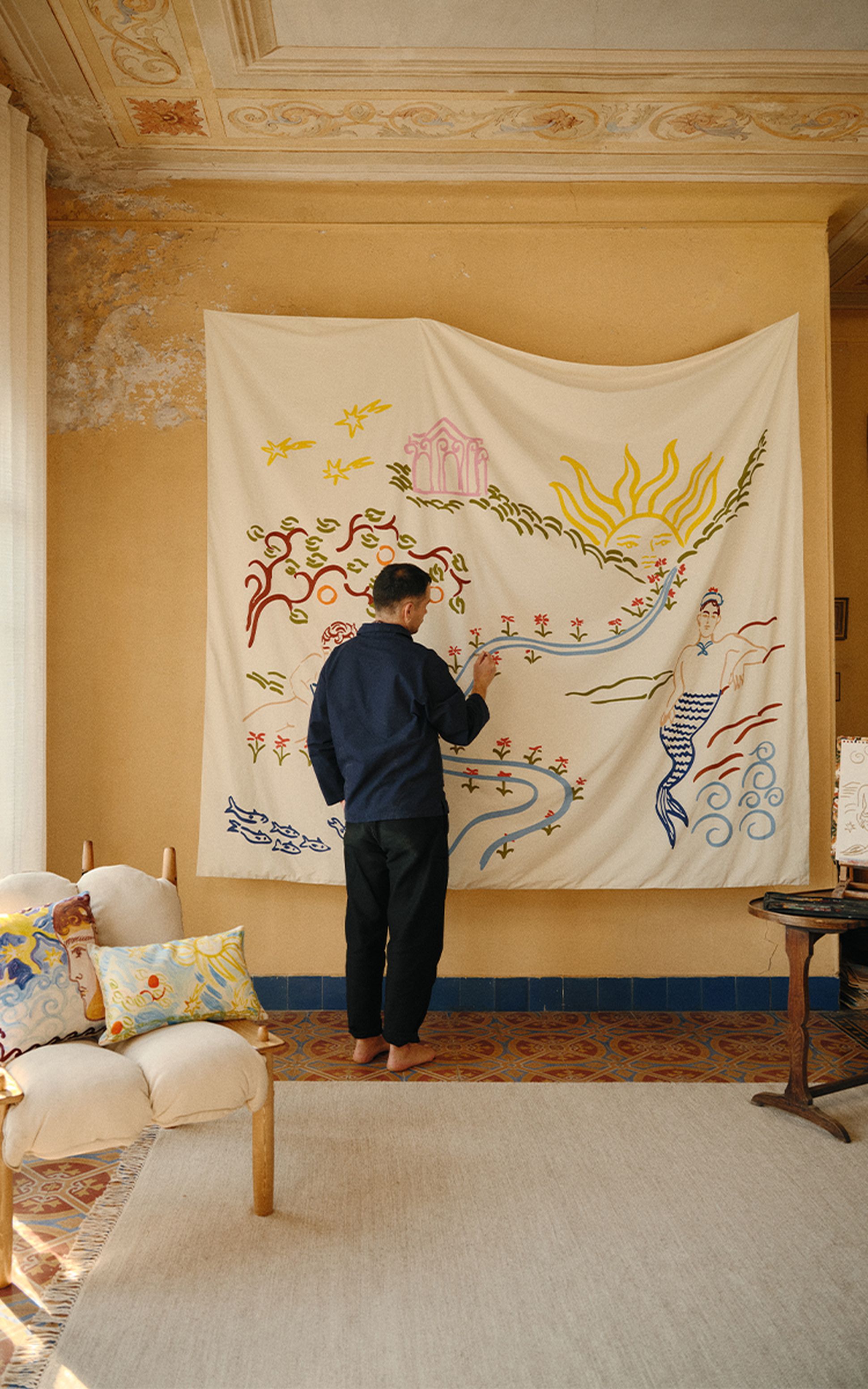 Une personne vêtue d'une chemise bleue peint sur une grande toile aux motifs fantaisistes, dans une pièce au sol à motifs et à la décoration vintage.