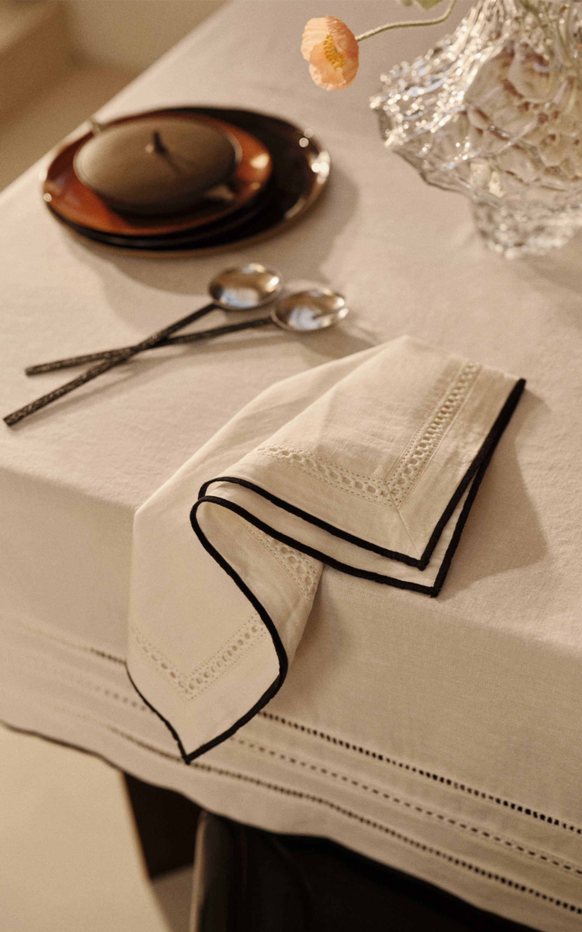 Une table élégamment dressée avec une serviette blanche brodée, deux cuillères en argent, un plat en céramique et une fleur de pêcher dans un vase en verre.