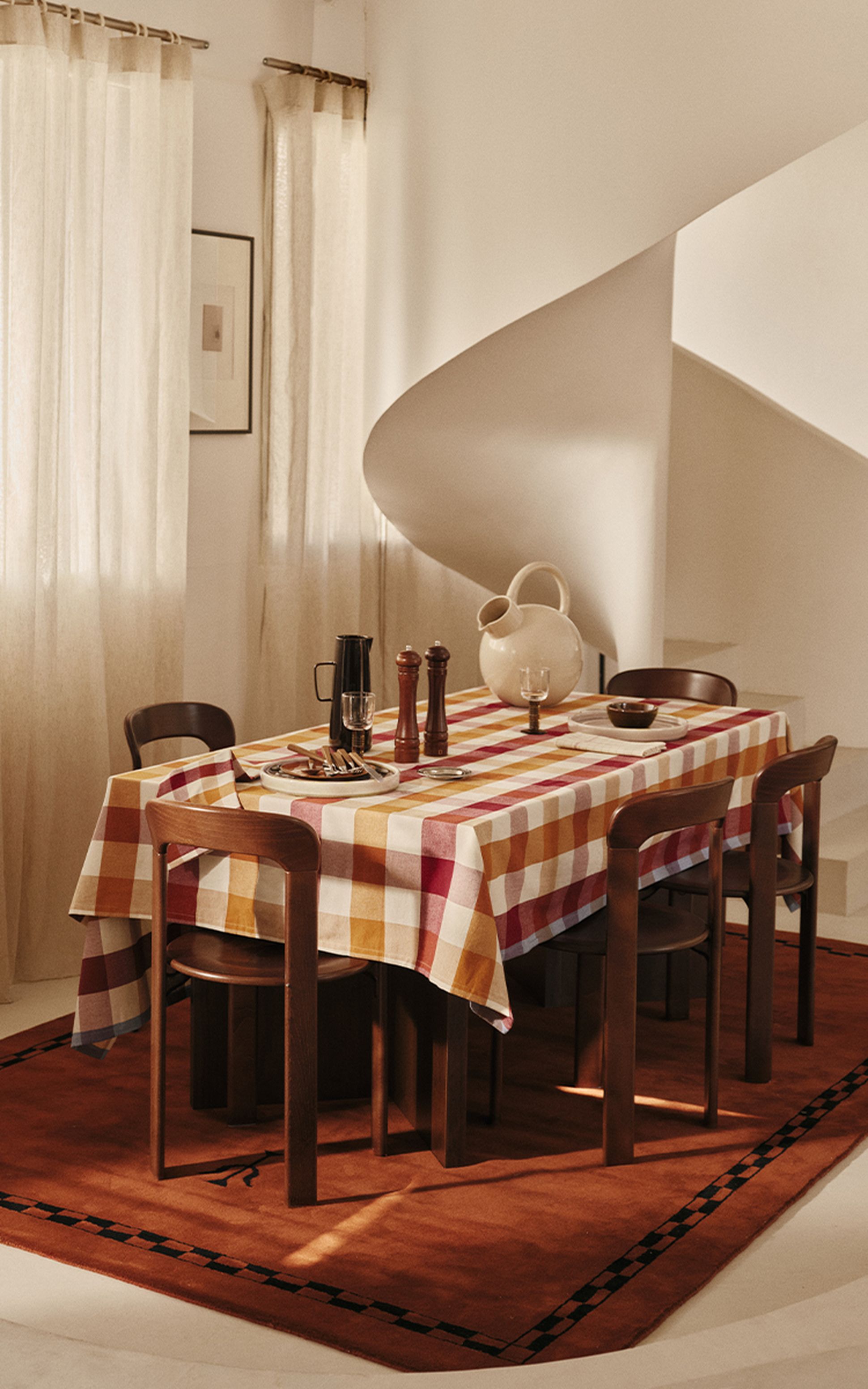 Salle à manger avec une table recouverte d'une nappe à carreaux, dressée avec une cruche, des salières et poivrières, et entourée de chaises en bois.