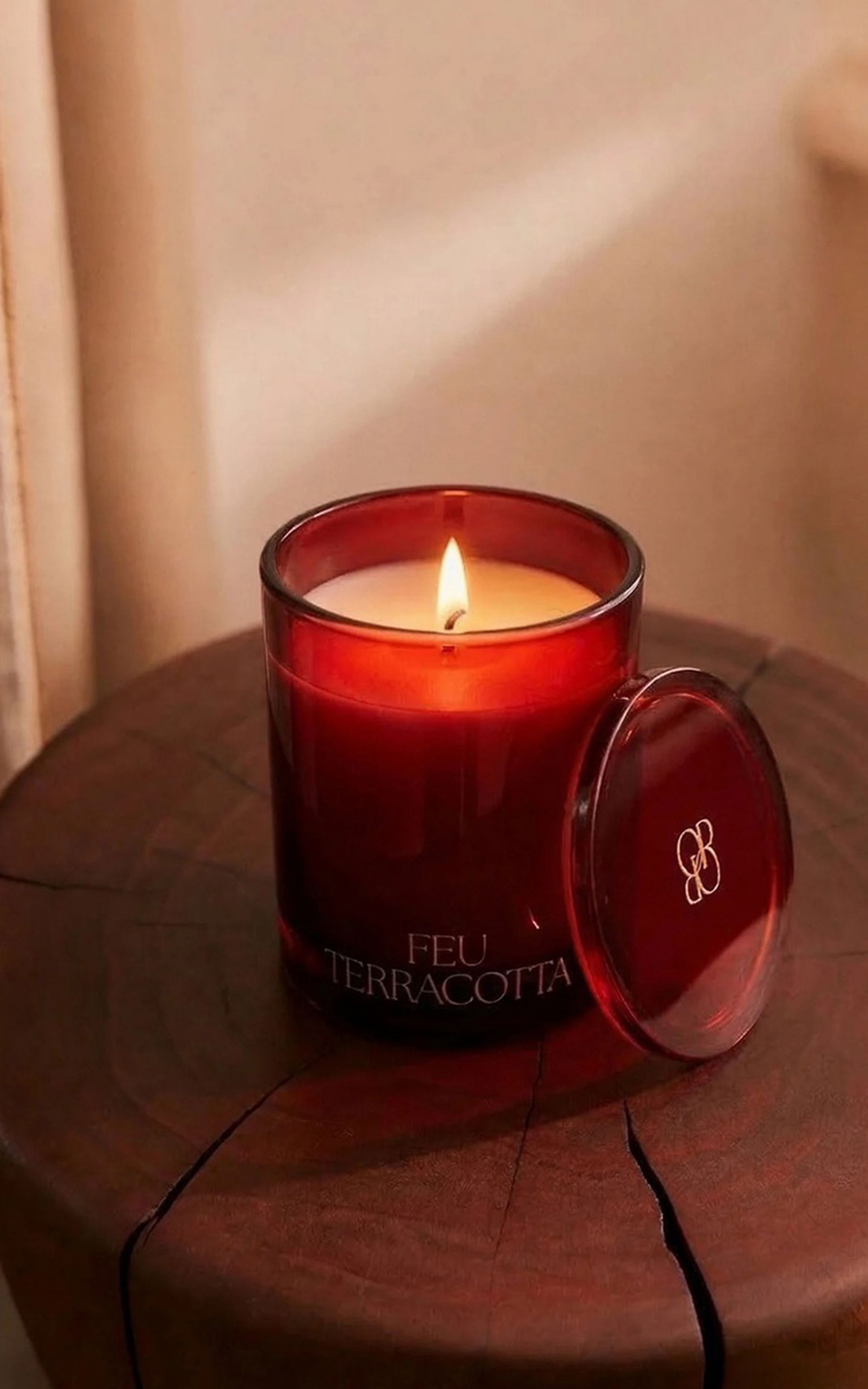 Une bougie allumée dans un récipient en verre rouge portant l'inscription « FEU TERRACOTTA », posée sur une surface en bois, son couvercle posé à côté.