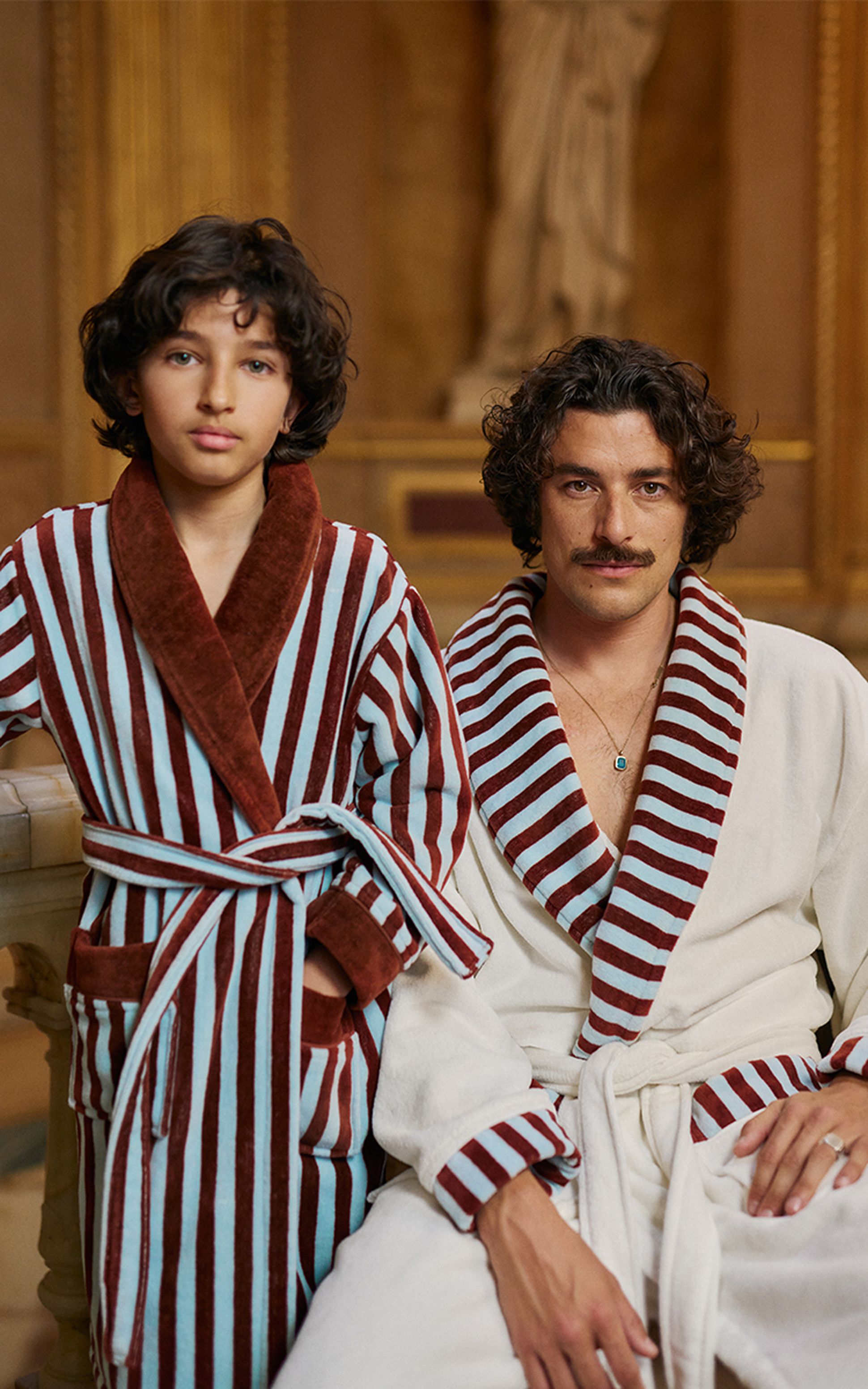 Un enfant et un adulte vêtus de peignoirs rayés assortis, l'un rouge et bleu, l'autre rouge et blanc, assis dans une pièce richement décorée avec une statue en arrière-plan.