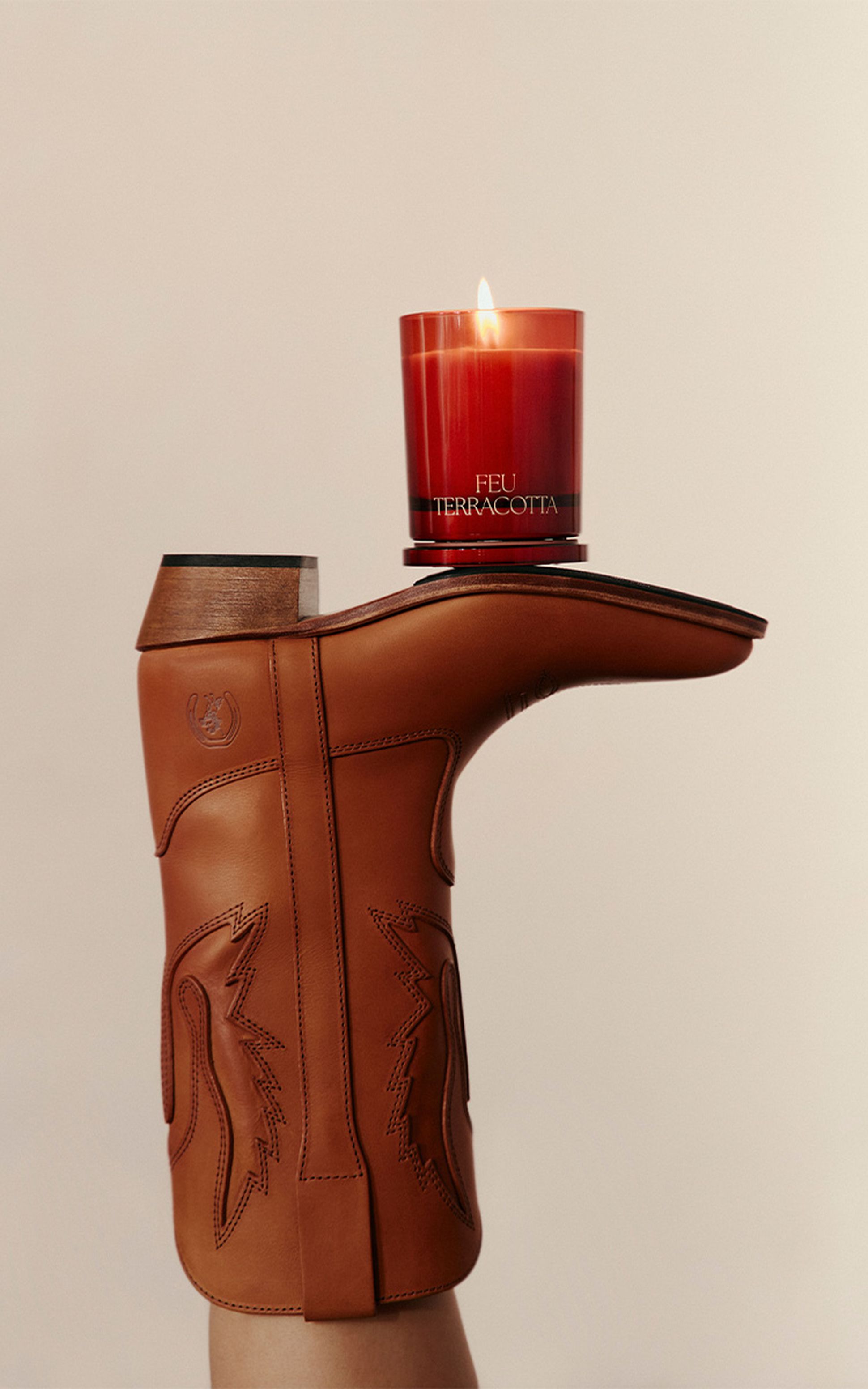 Une botte de cow-boy marron à l'envers, avec une bougie rouge allumée portant l'inscription « FEU TERRACOTTA » en équilibre sur sa semelle, sur un fond uni.