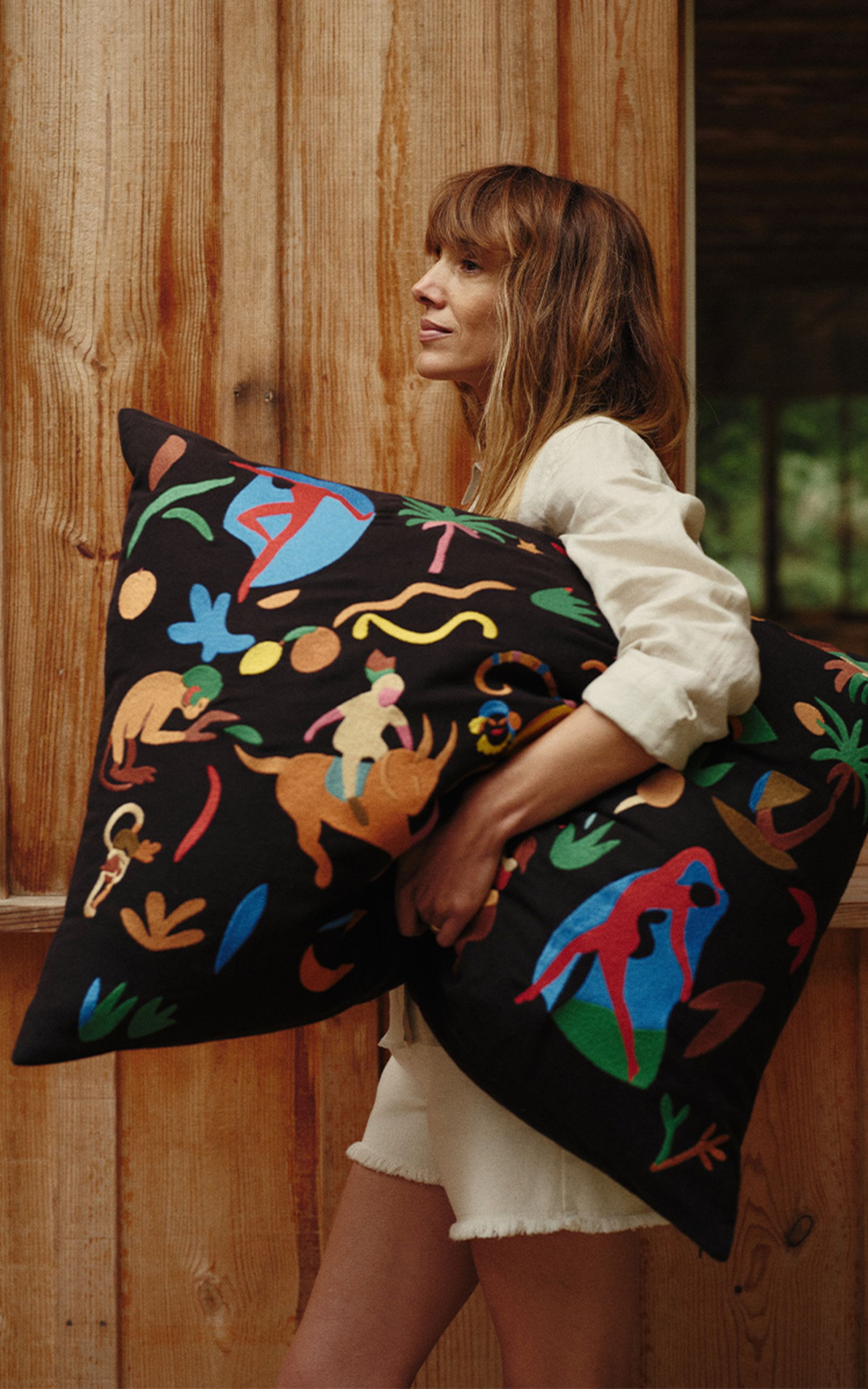Une femme vêtue d'une tenue blanche tient un grand coussin coloré à motifs de personnages et de formes, debout devant un mur en bois.