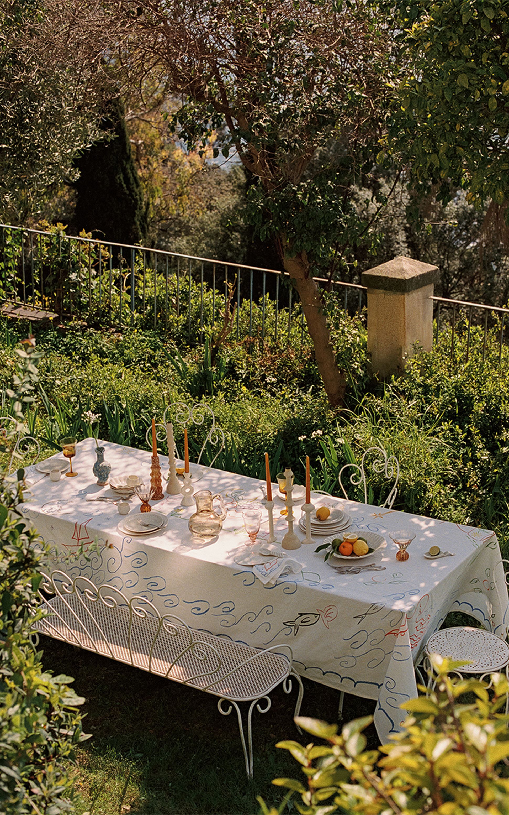 Une table d'extérieur dressée pour un repas, avec une nappe blanche, des bougies et des fruits, entourée d'une végétation luxuriante et à l'ombre d'un arbre.