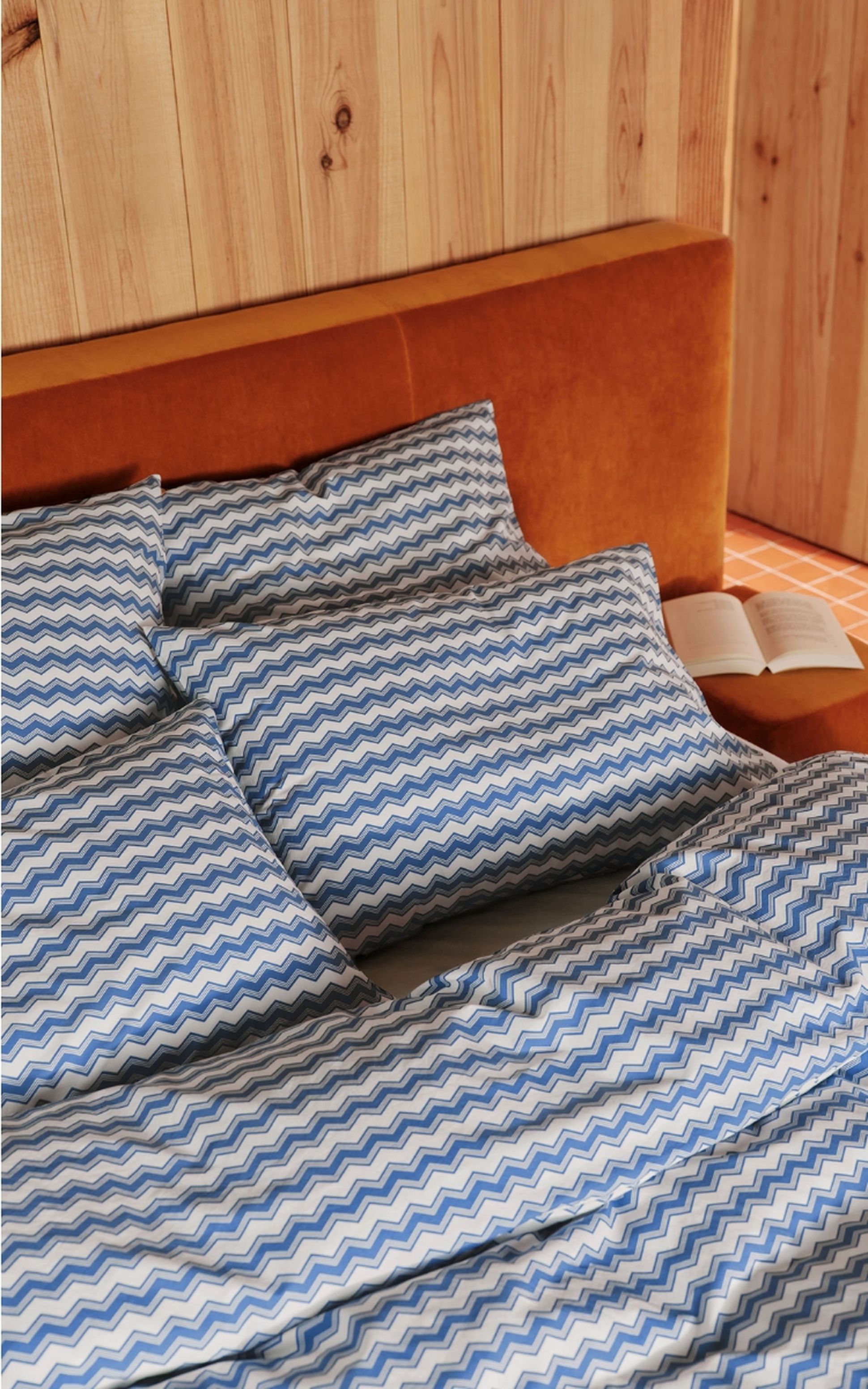 Une parure de lit bleue et blanche à motifs en chevrons sur un lit à tête de lit marron, adossé à un mur en bois, avec un livre ouvert à proximité.