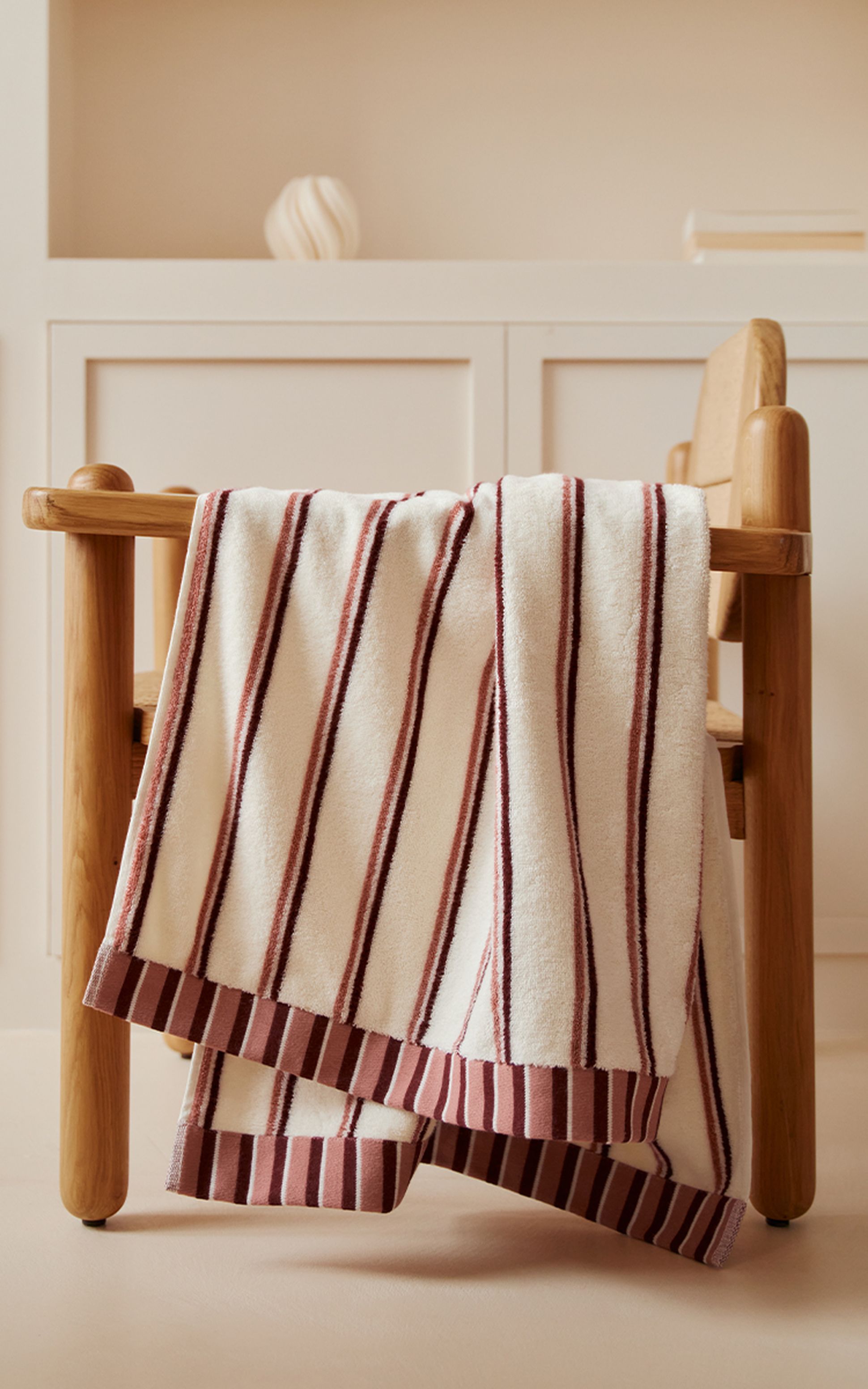 Une serviette blanche à rayures bordeaux est posée sur une chaise en bois dans une pièce minimaliste aux murs beiges, avec une étagère en arrière-plan.