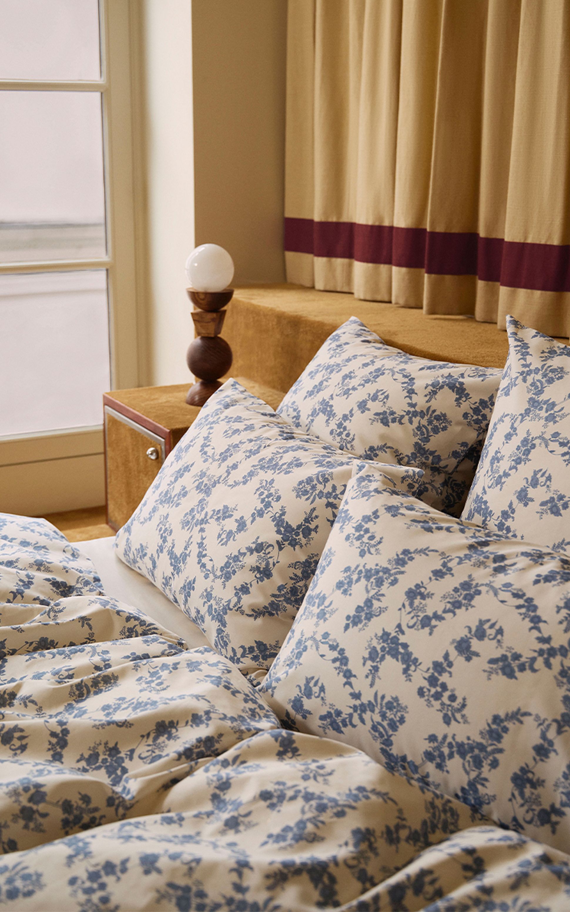 Chambre à coucher avec un lit soigneusement fait, une literie à motifs floraux bleus, un coussin décoratif marron, une grande fenêtre et des œuvres d'art accrochées au mur.