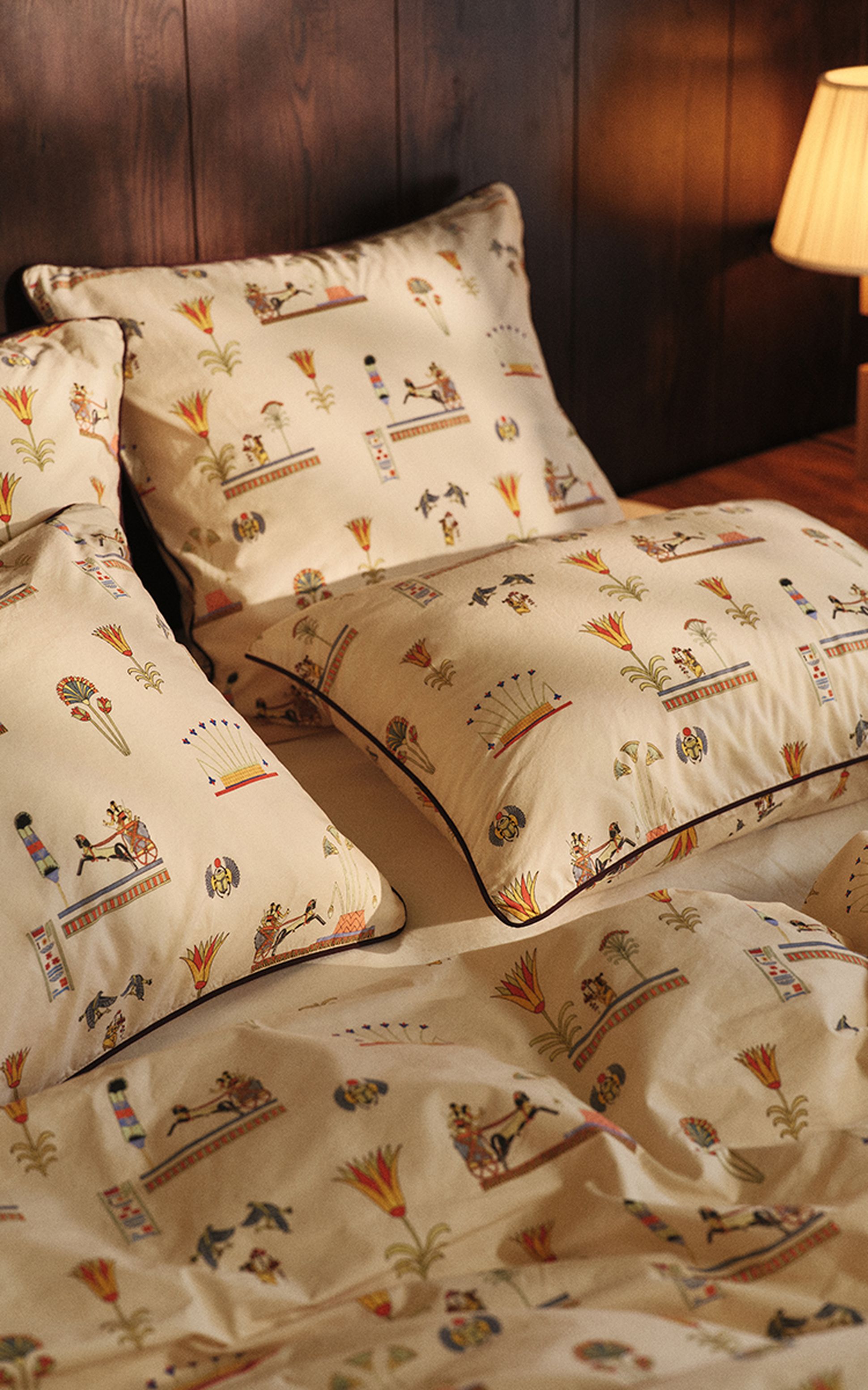 Cream-colored bedding with Egyptian-inspired patterns, featuring hieroglyphs and symbols, set against a dark wood paneled wall with a lamp.