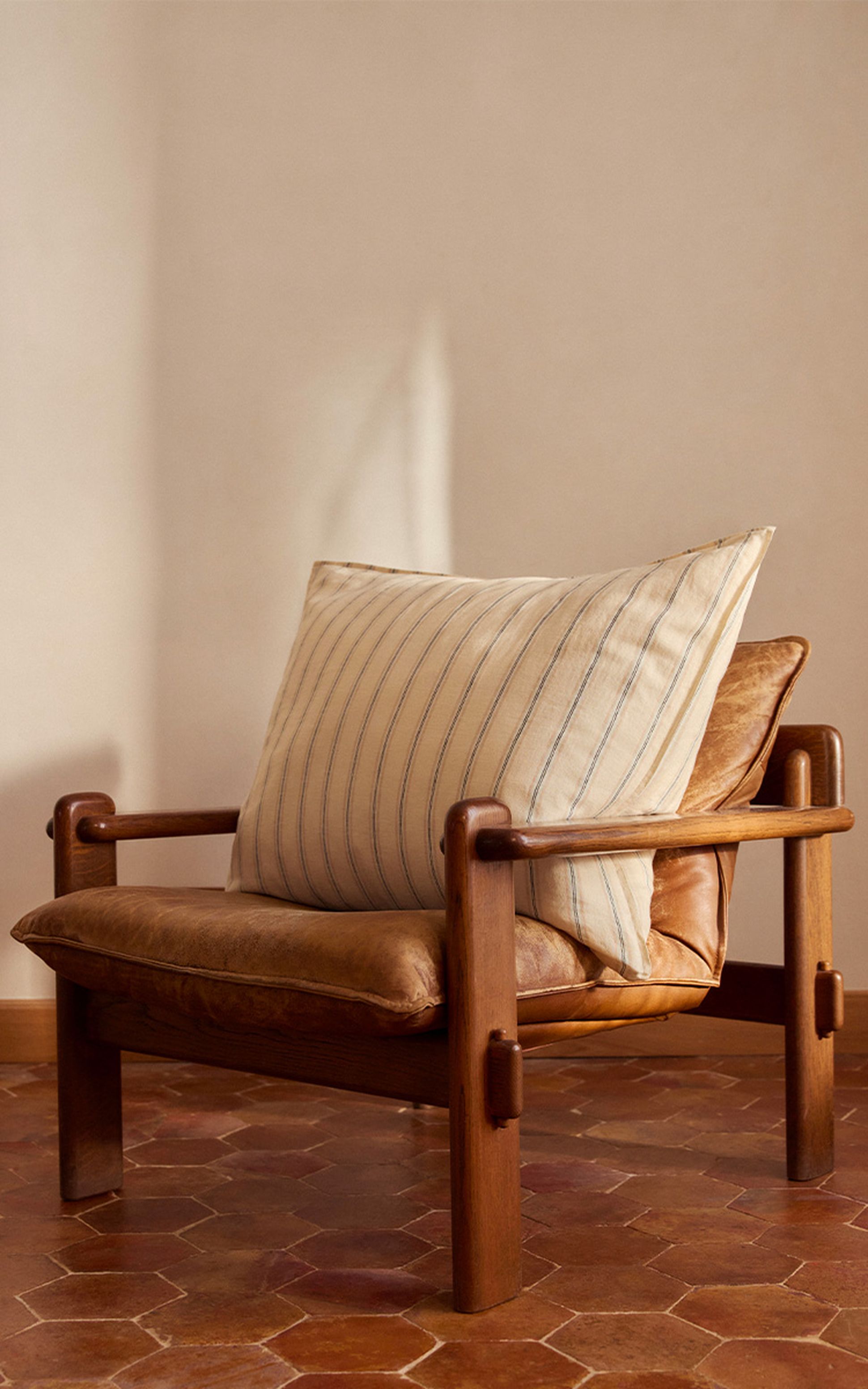 Un fauteuil en cuir orné d'un coussin beige rayé, posé sur un sol carrelé de couleur terre cuite, devant un mur de couleur neutre.
