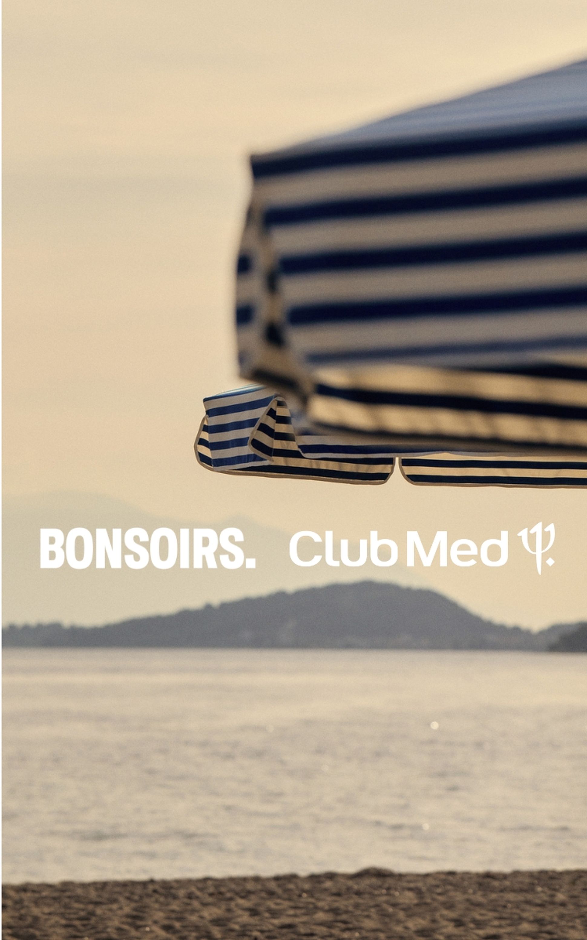 Des parasols rayés au bord d'une mer calme, avec les montagnes en arrière-plan, sur lesquels figure l'inscription « BONSOIRS. Club Med. »