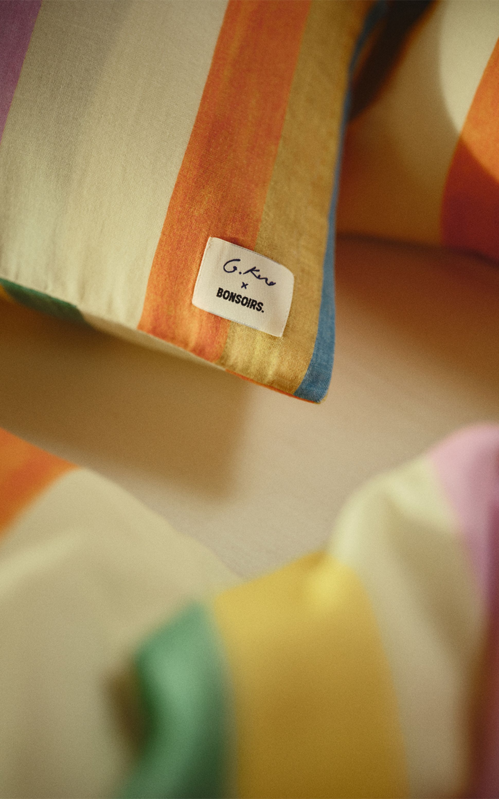 Gros plan sur des coussins rayés colorés portant une étiquette sur laquelle on peut lire « G. Kero x BONSOIRS ». Le tissu est composé de rayures orange, jaunes, bleues, vertes et roses.