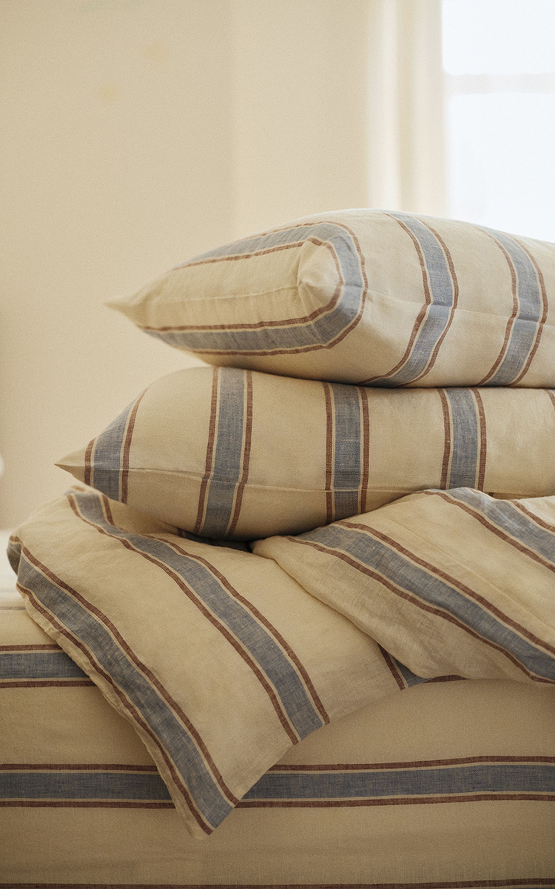 Une pile de coussins beiges à rayures bleues et marron posés sur un couvre-lit assorti, dans une pièce baignée d'une lumière tamisée.