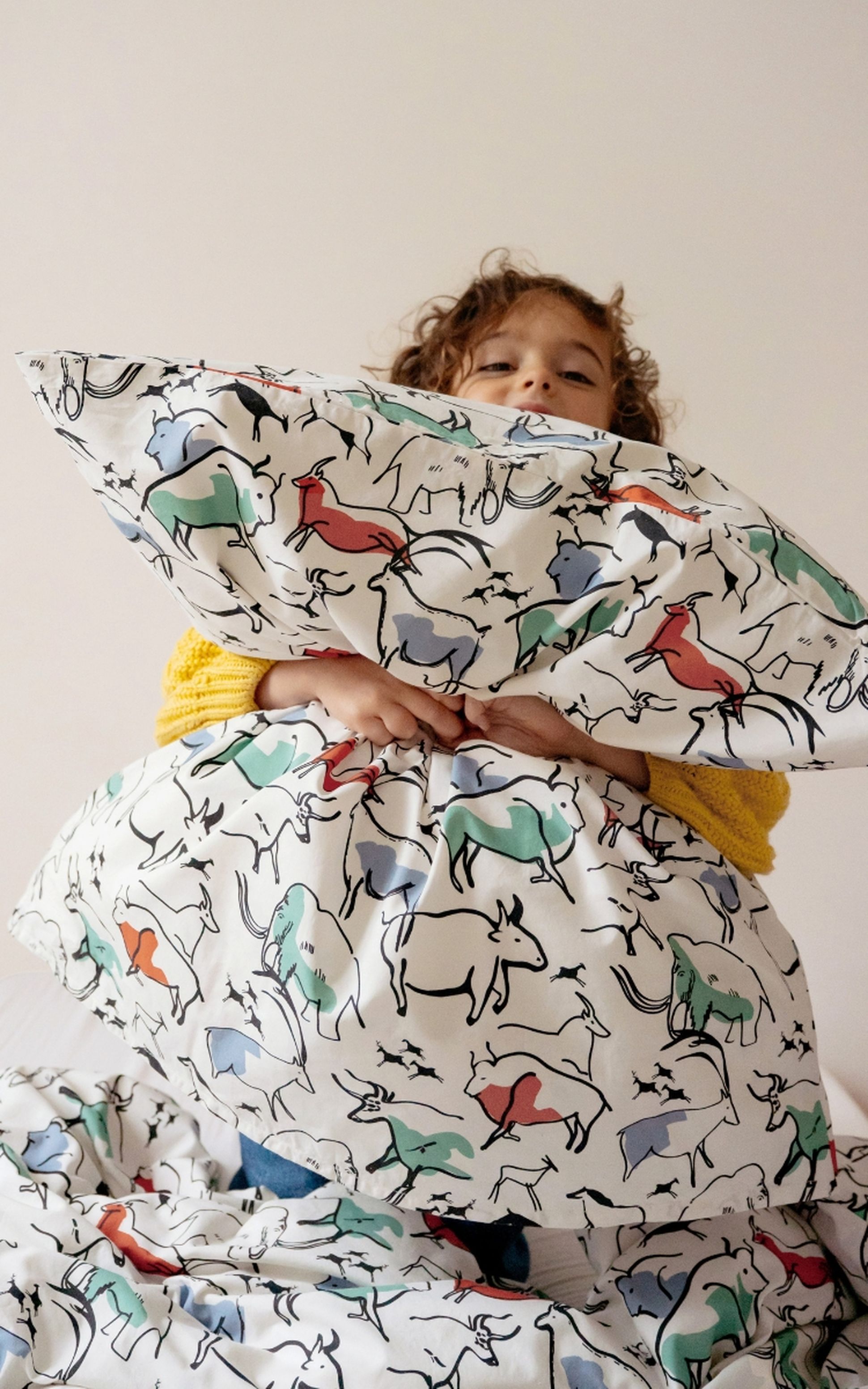 Enfant serrant dans ses bras un oreiller blanc orné de dessins d'animaux colorés, vêtu d'un pull jaune, sur fond uni.