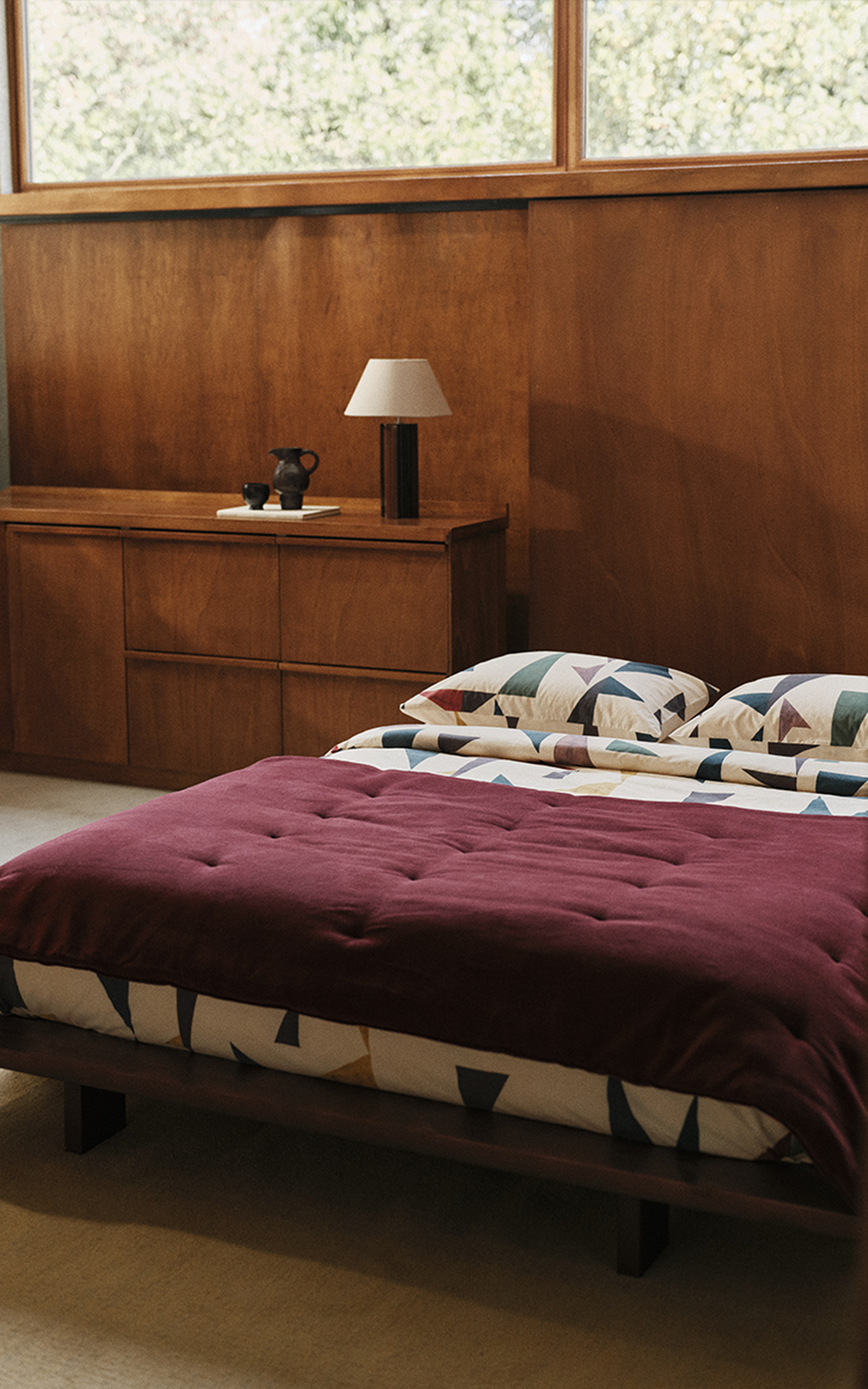 Chambre à coucher avec une commode en bois, une lampe de chevet, un lit recouvert d'une couette bordeaux et des coussins aux motifs géométriques, le tout devant un mur lambrissé.