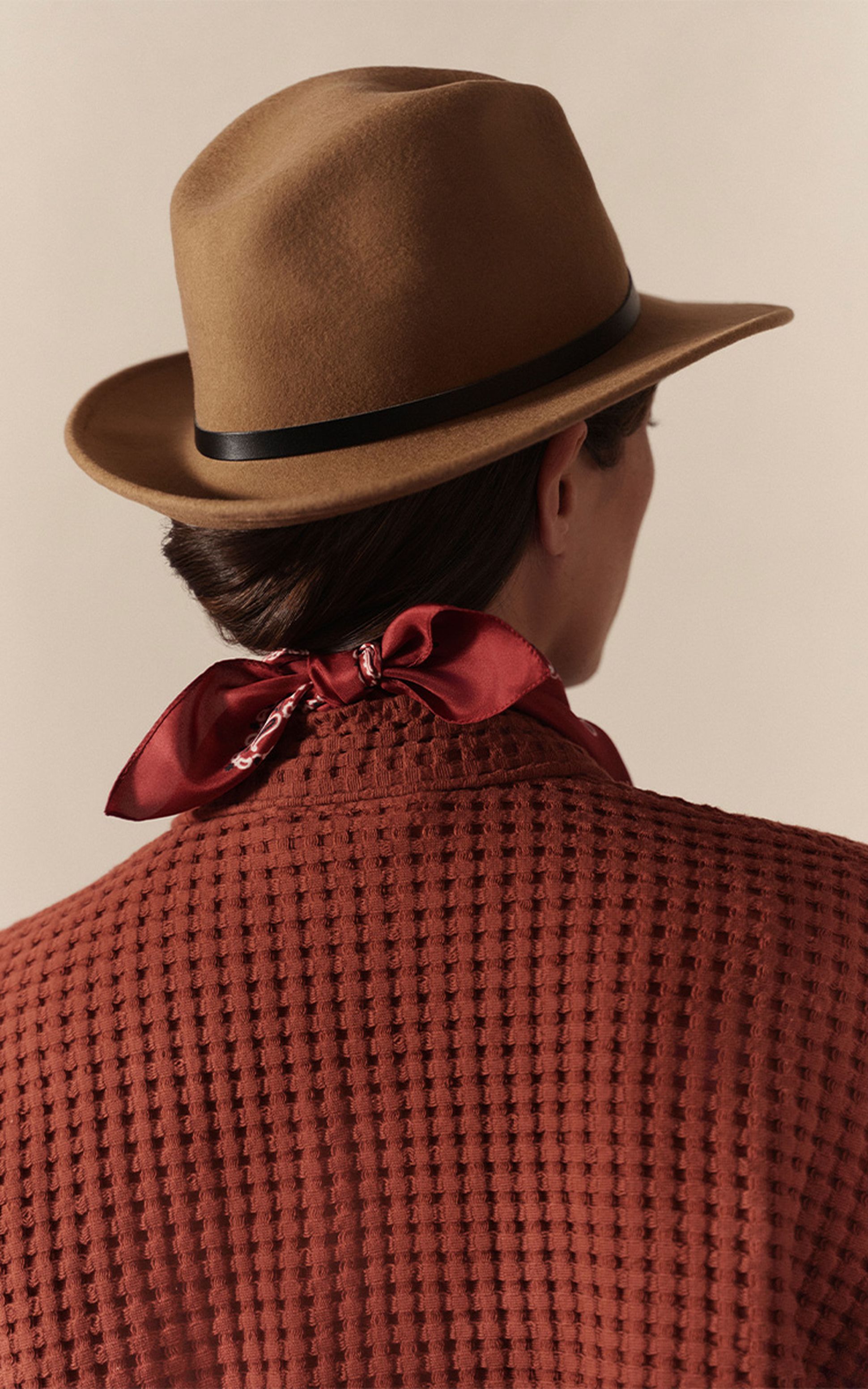 Personne portant un chapeau marron et un peignoir marron texturé, avec une écharpe rouge nouée autour du cou, dos à la caméra, devant un fond neutre.