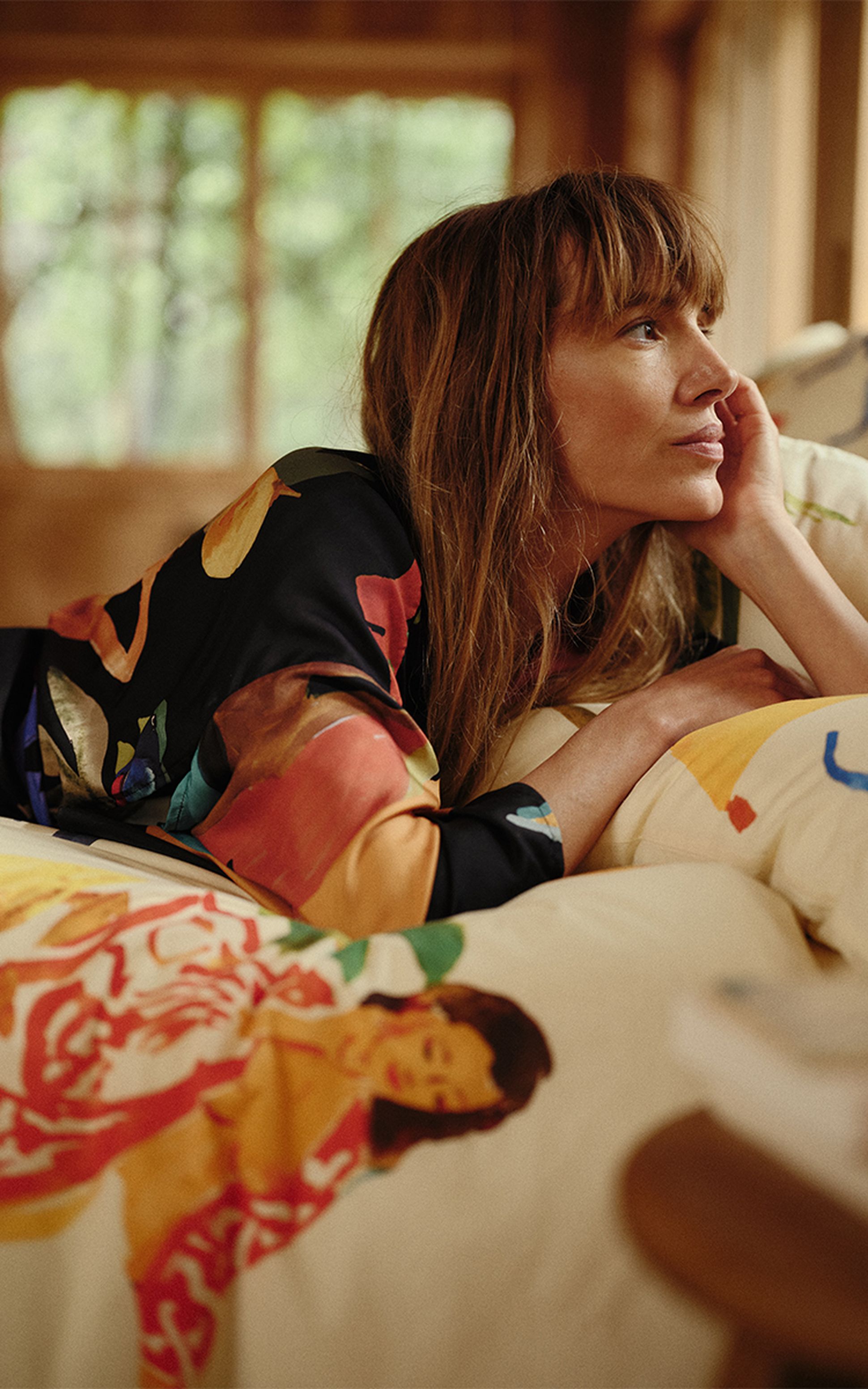 Une femme aux cheveux longs, vêtue d'une robe colorée, est allongée sur un coussin à motifs et regarde pensivement à travers une fenêtre floue en arrière-plan.