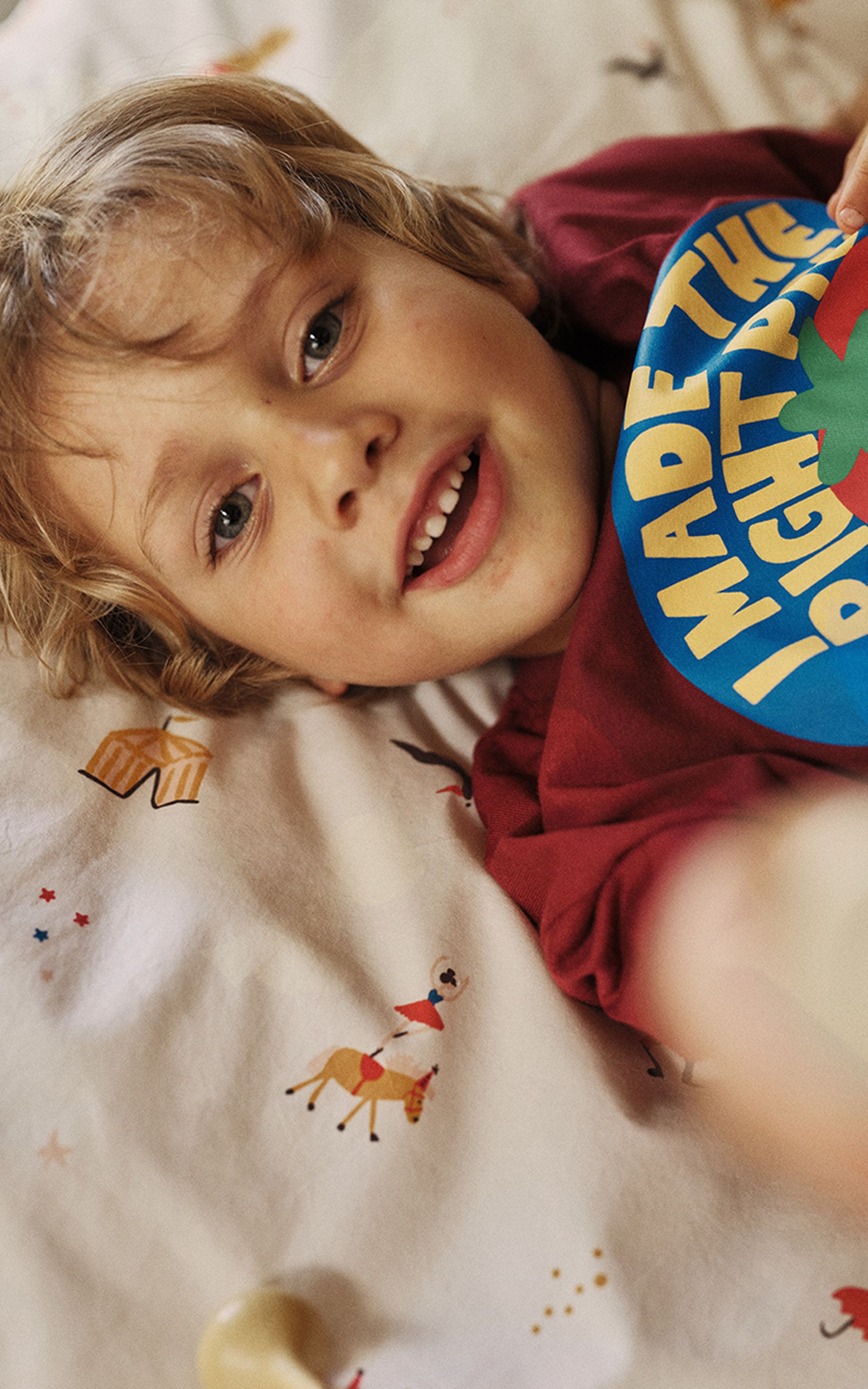Un enfant souriant aux cheveux blonds est allongé sur un lit recouvert d'un drap coloré et fantaisiste. Il porte un t-shirt rouge orné d'une inscription jaune.