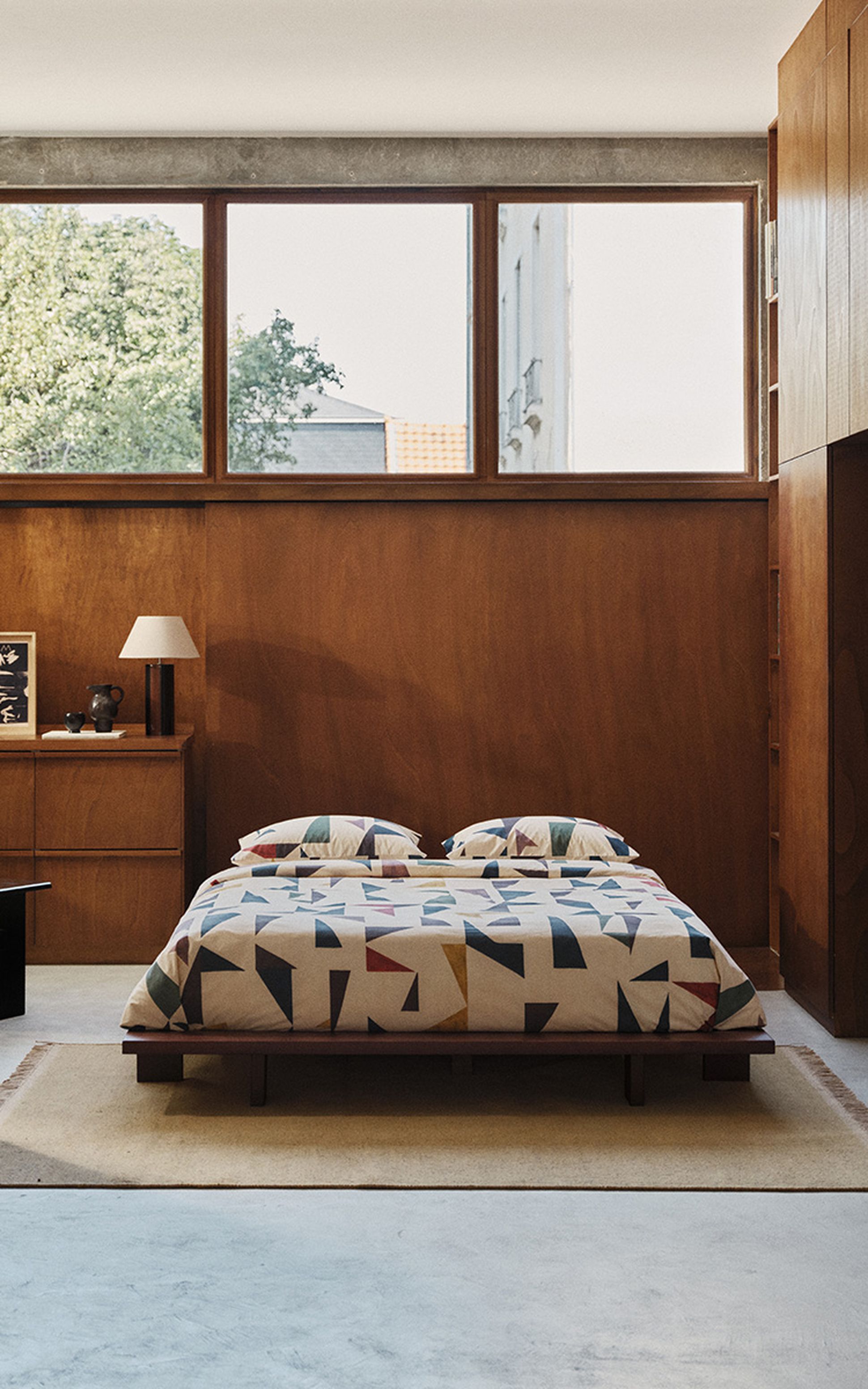 Une chambre douillette avec un lit bas recouvert d'une parure aux motifs géométriques colorés, des murs en bois et de grandes fenêtres donnant sur les arbres à l'extérieur.