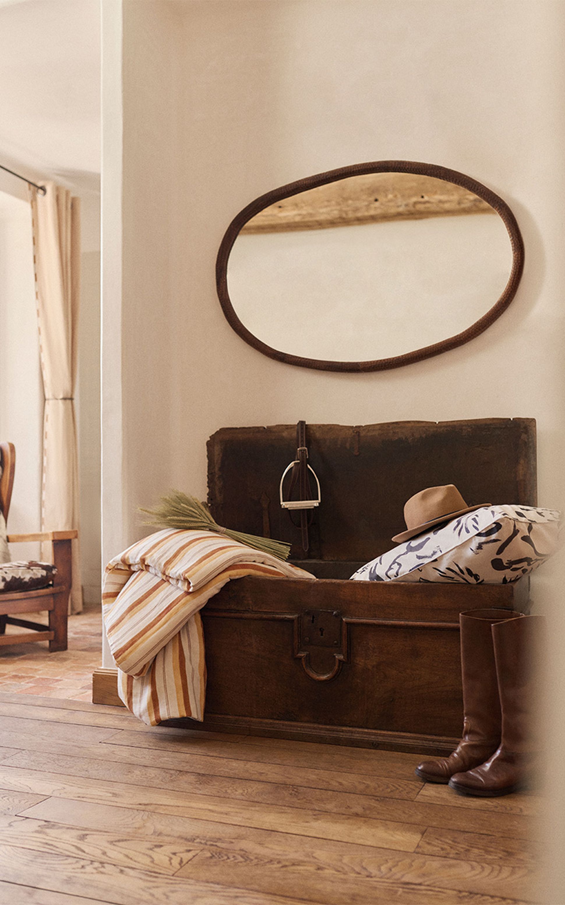 Une malle rustique en bois contenant des coussins, un chapeau et des bottes est placée sous un miroir ovale dans une pièce chaleureusement éclairée avec un sol en bois.