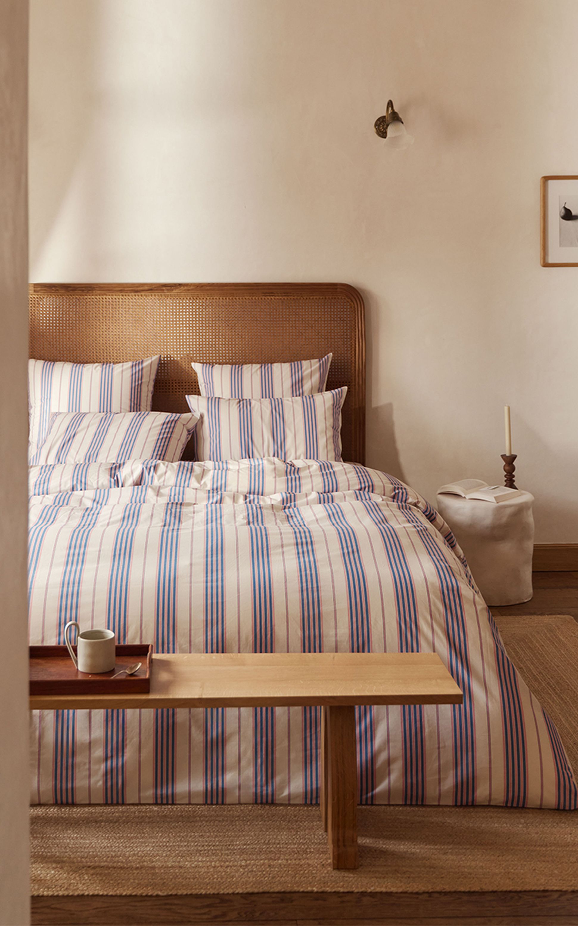 Chambre confortable avec une tête de lit en rotin et un lit soigneusement fait, recouvert d'une literie à rayures bleues. Un banc en bois et un petit plateau avec une tasse se trouvent devant.