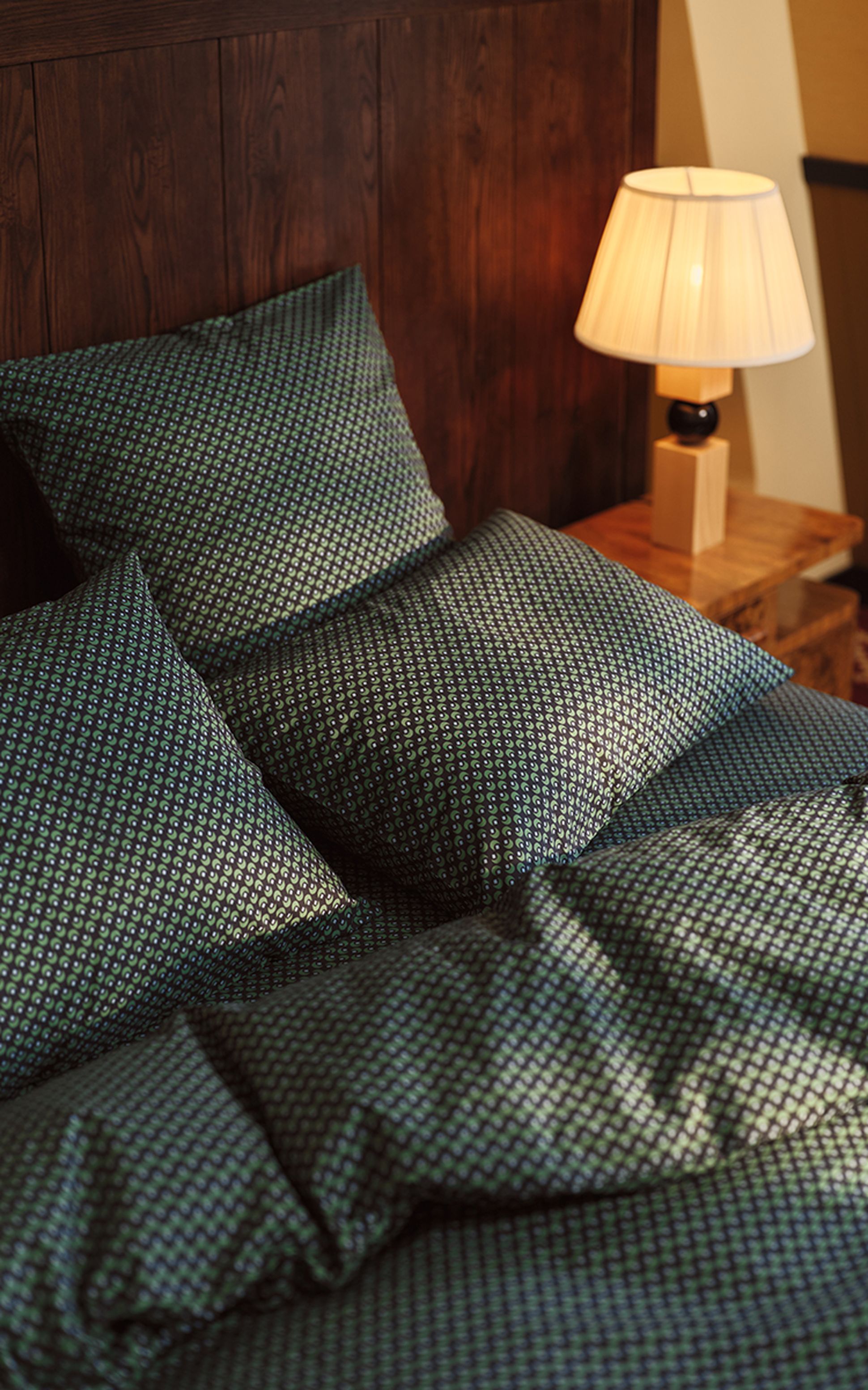 Bed with green patterned sheets and pillows beside a wooden nightstand with a lit lamp.