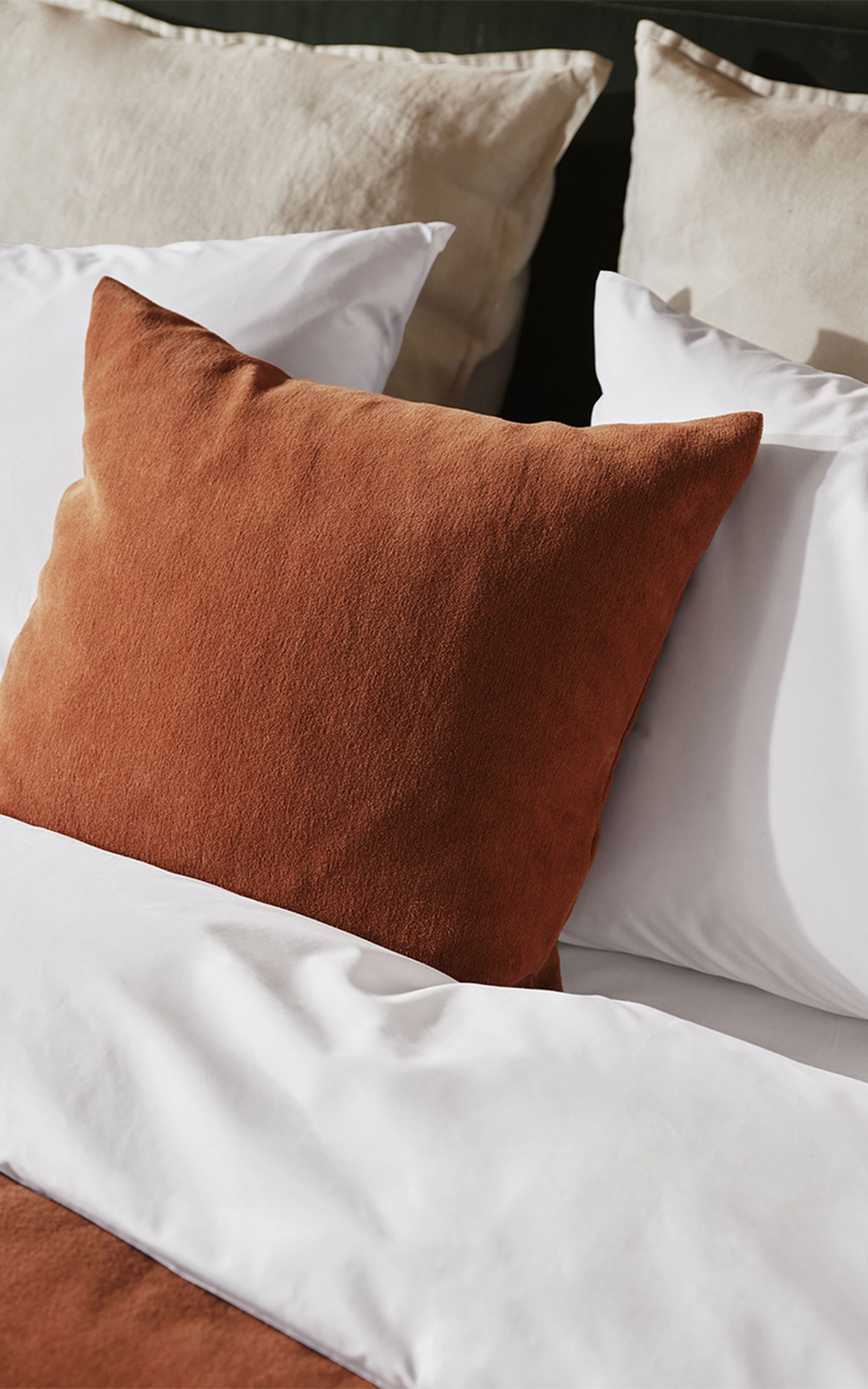 Un lit bien fait, avec un coussin décoratif couleur rouille, des draps blancs et des oreillers beiges en arrière-plan.