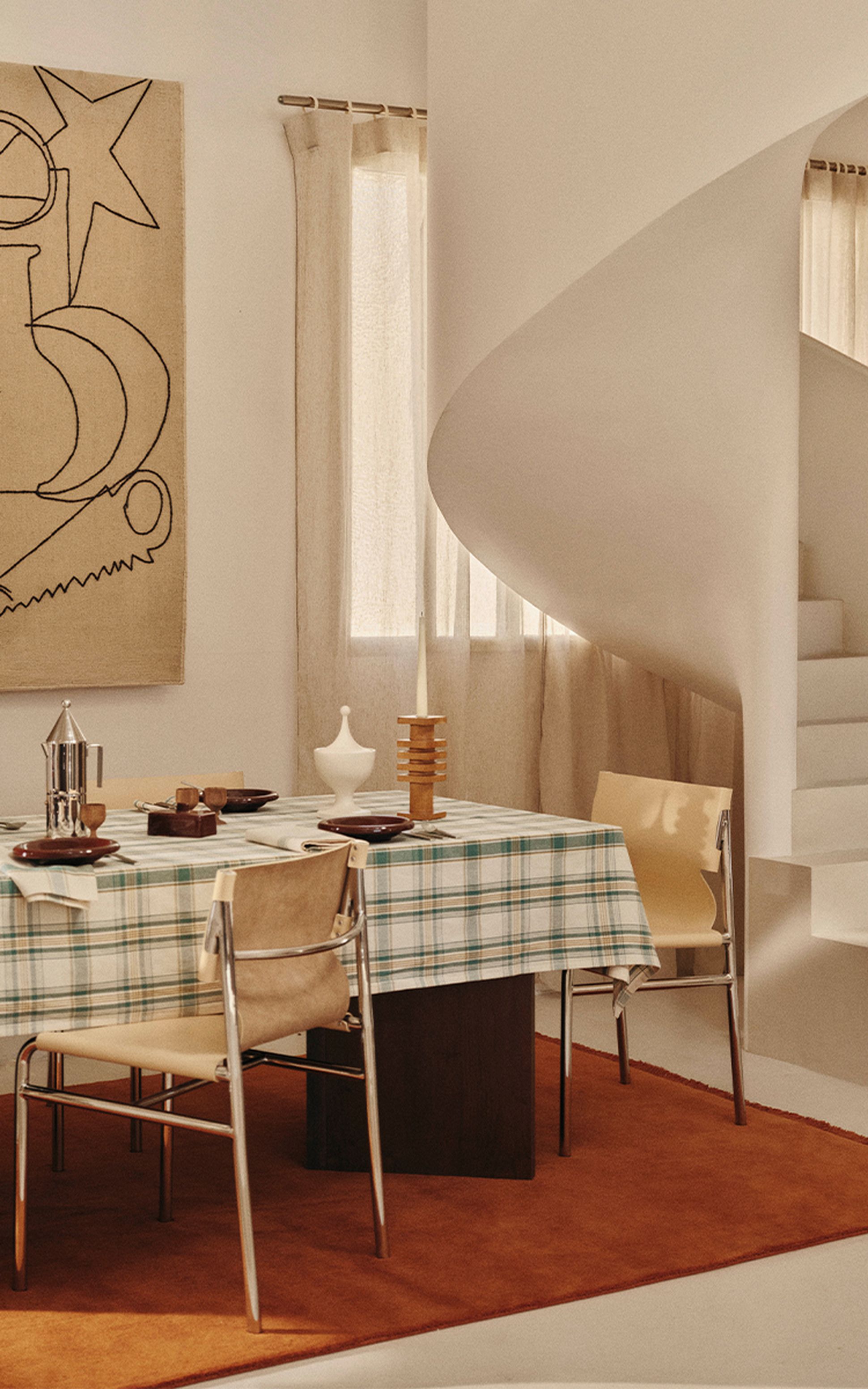Une salle à manger moderne avec une nappe à carreaux, des chaises minimalistes, des œuvres d'art abstraites aux murs et un escalier en colimaçon. Des tons neutres et chaleureux imprègnent l'espace.
