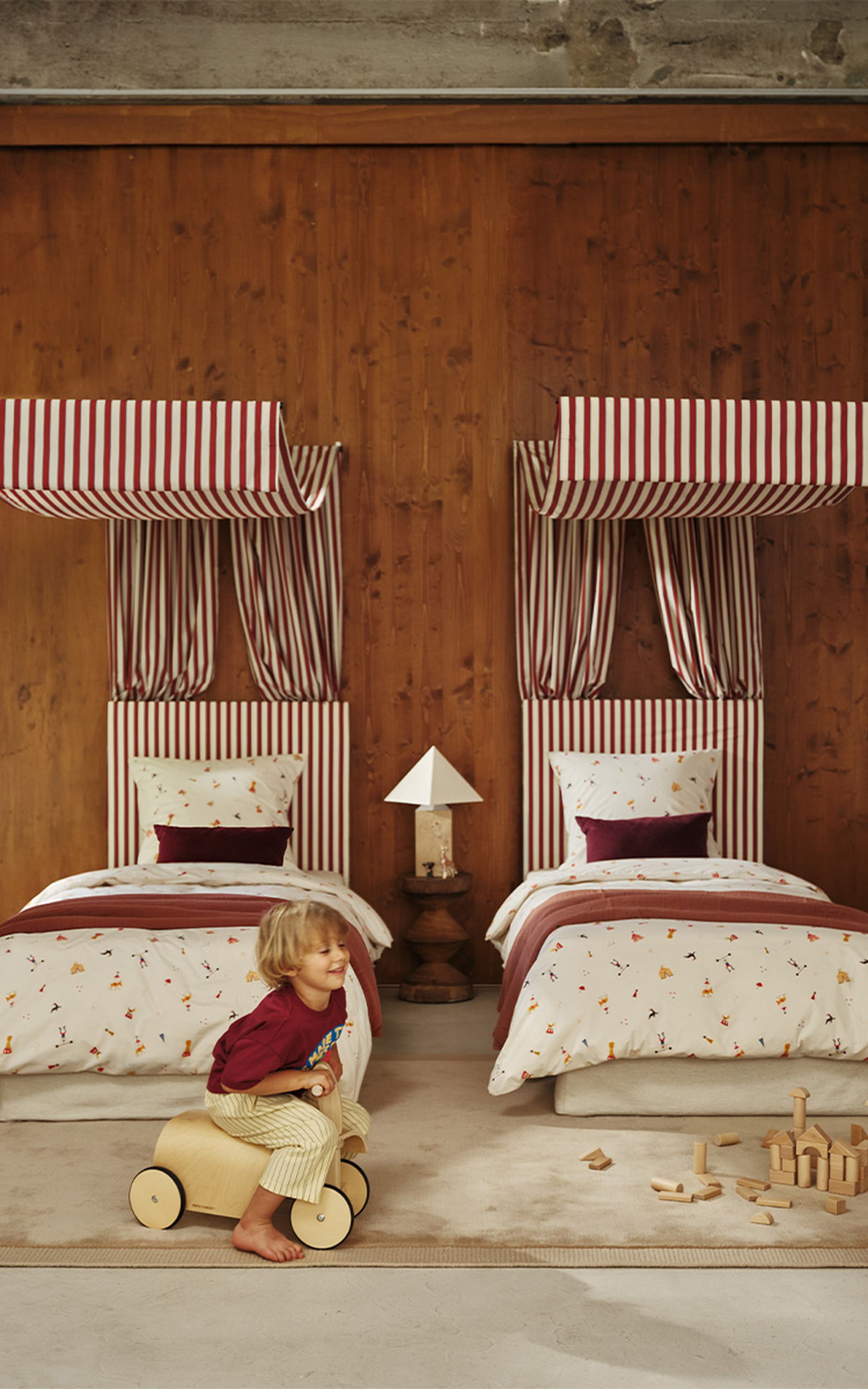Un enfant joue avec un véhicule-jouet en bois dans une chambre chaleureuse dotée de deux lits, de têtes de lit à rayures, d'une literie rouge et blanche et de jouets en bois éparpillés sur le sol.