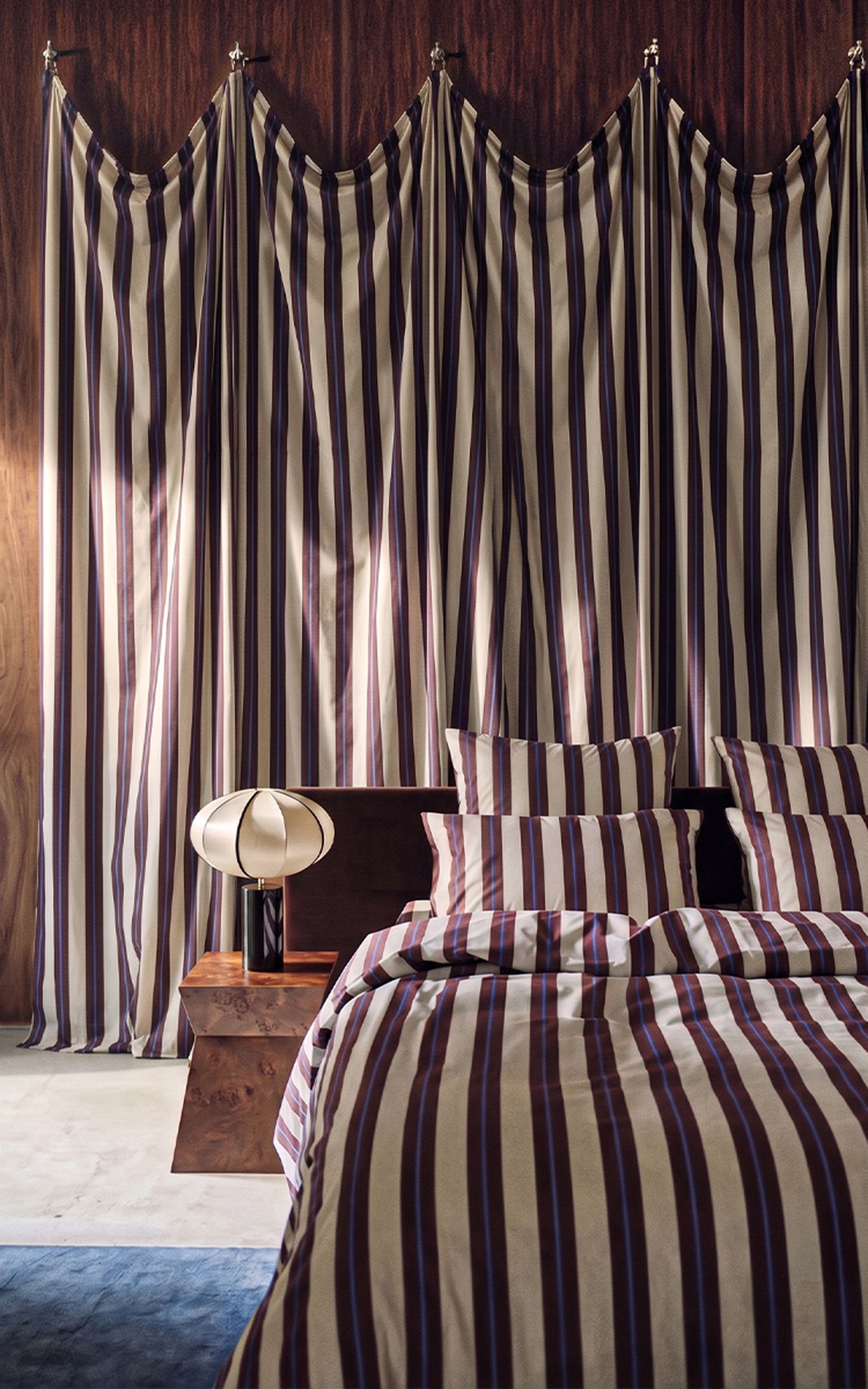 Une chambre avec une parure de lit à rayures marron et crème et des rideaux assortis, une table de chevet en bois et une lampe décorative.