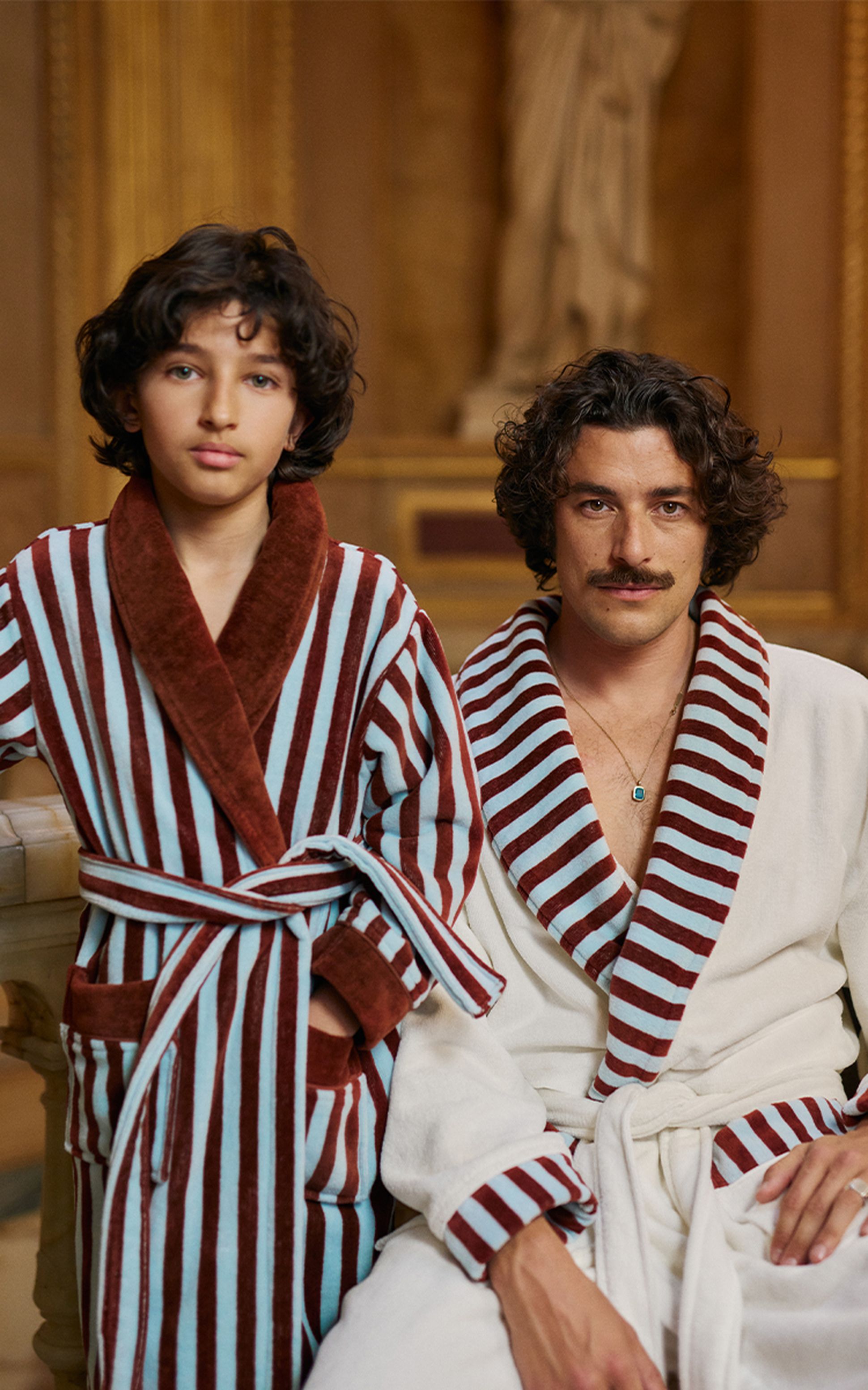 Deux personnes vêtues de robes rayées, l'une assise, l'autre debout, toutes deux aux cheveux bouclés, dans une pièce avec un décor en arrière-plan.