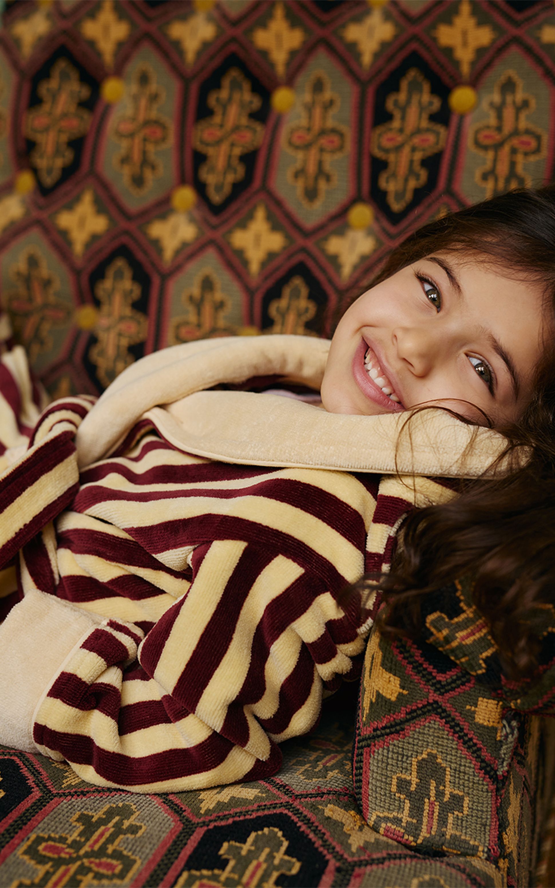 Un enfant souriant, vêtu d'un peignoir rayé bordeaux et beige, est allongé sur une chaise ornée d'un motif géométrique.