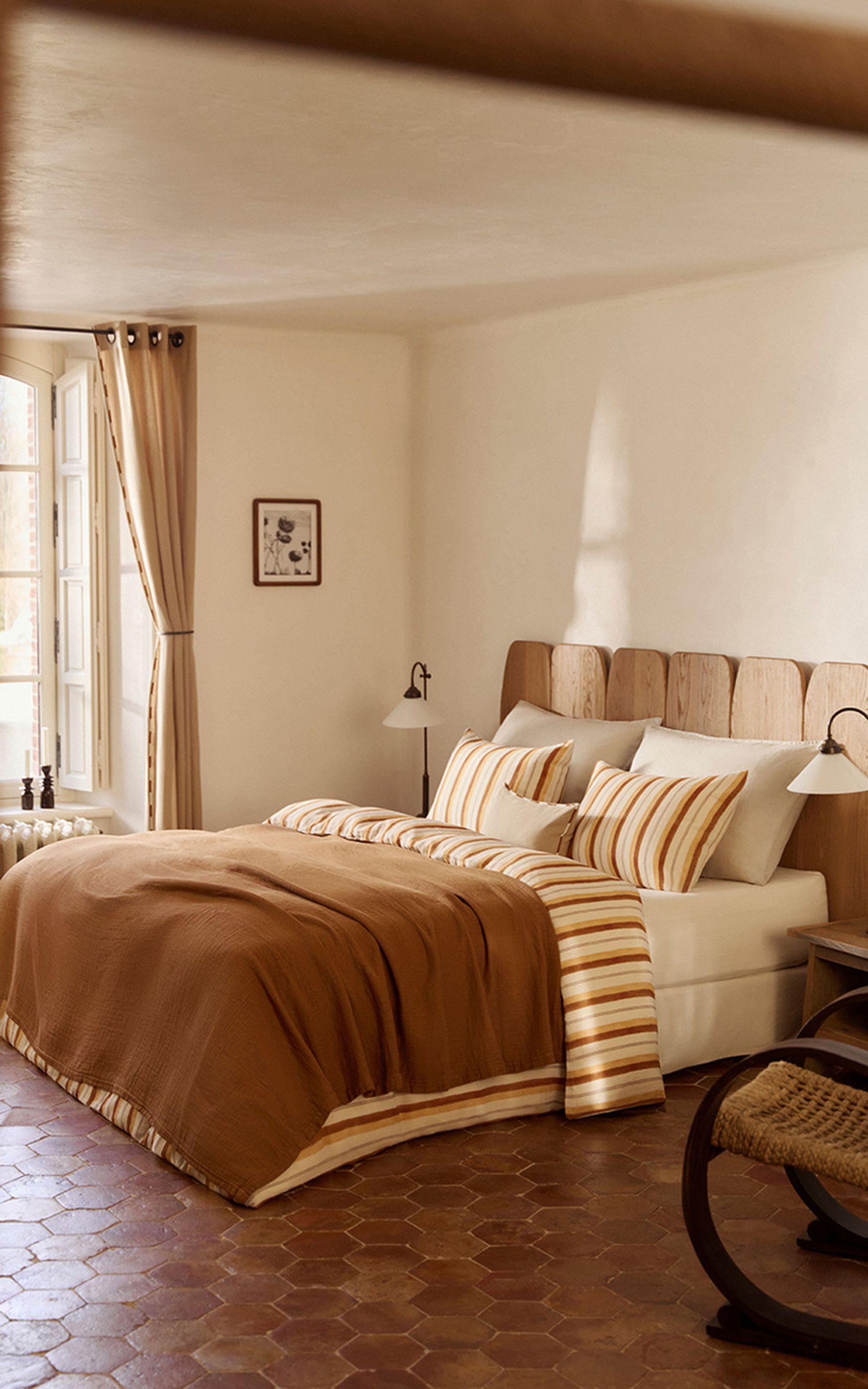 Chambre confortable avec une tête de lit en bois, des oreillers rayés et un couvre-lit marron. L'éclairage chaleureux et la lampe de chevet créent une ambiance paisible.