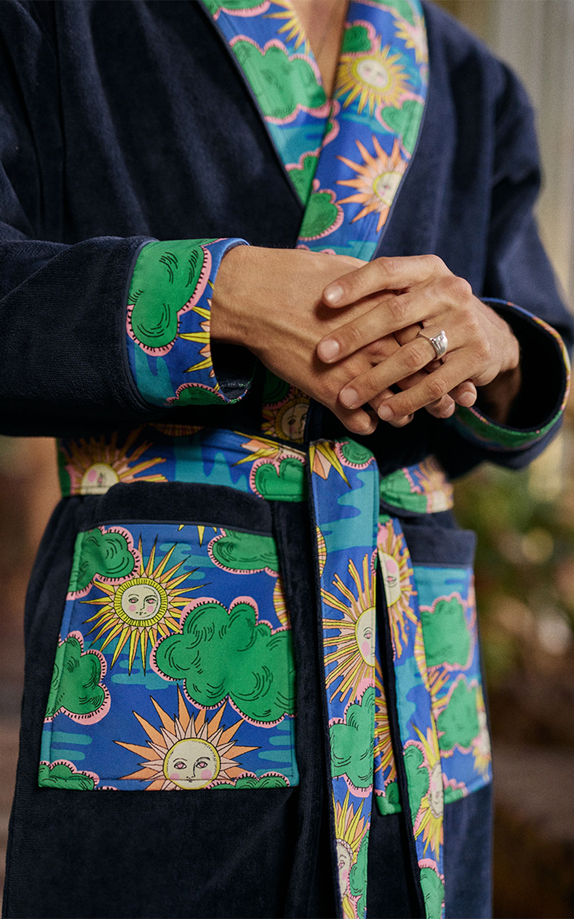 Personne vêtue d'un peignoir sombre orné de motifs colorés représentant des soleils et des nuages, les mains jointes, portant une bague.