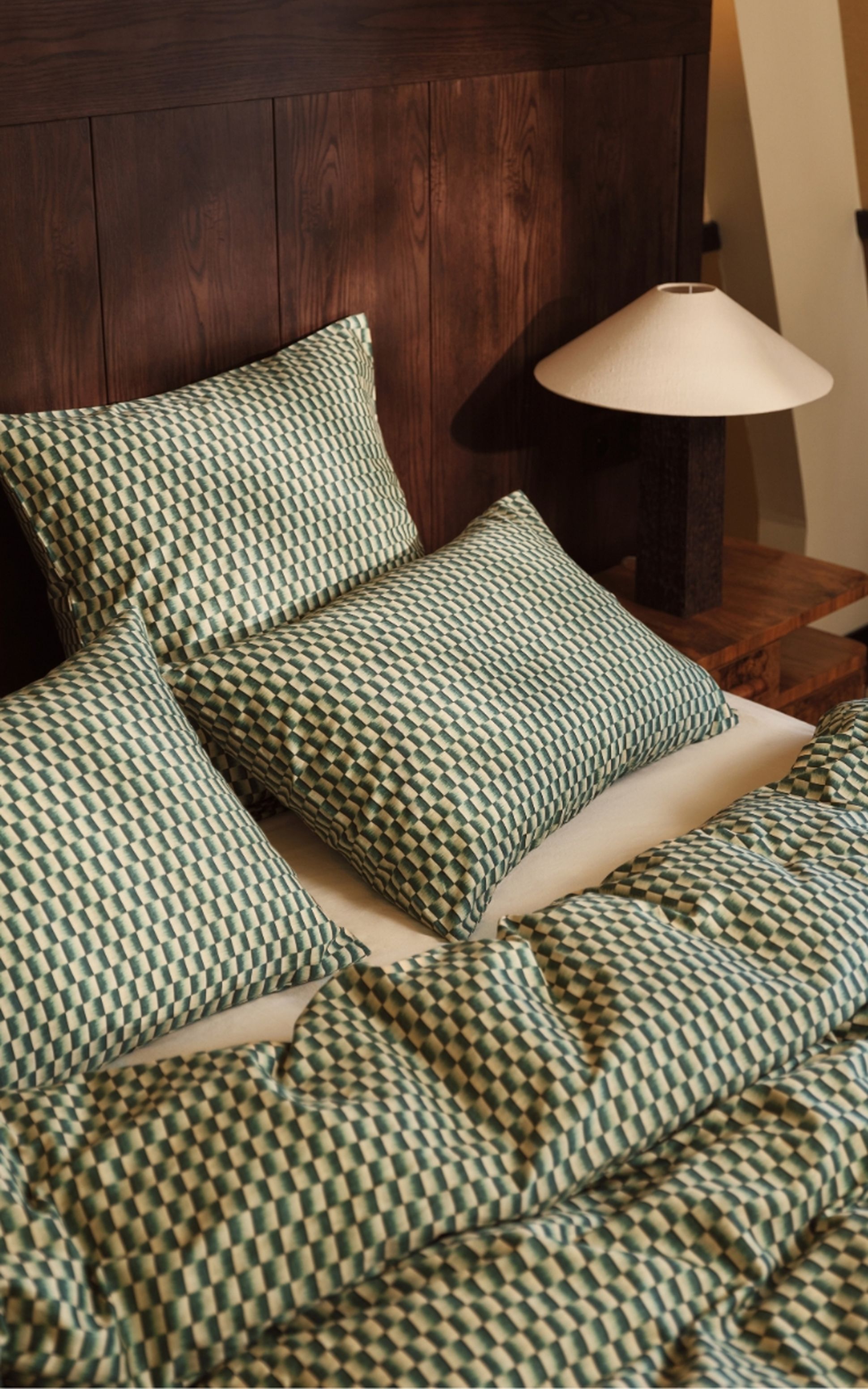 Un lit avec une parure de lit et des oreillers à motifs carreaux verts et beiges, à côté d'une tête de lit en bois et d'une table de chevet sur laquelle est posée une lampe.
