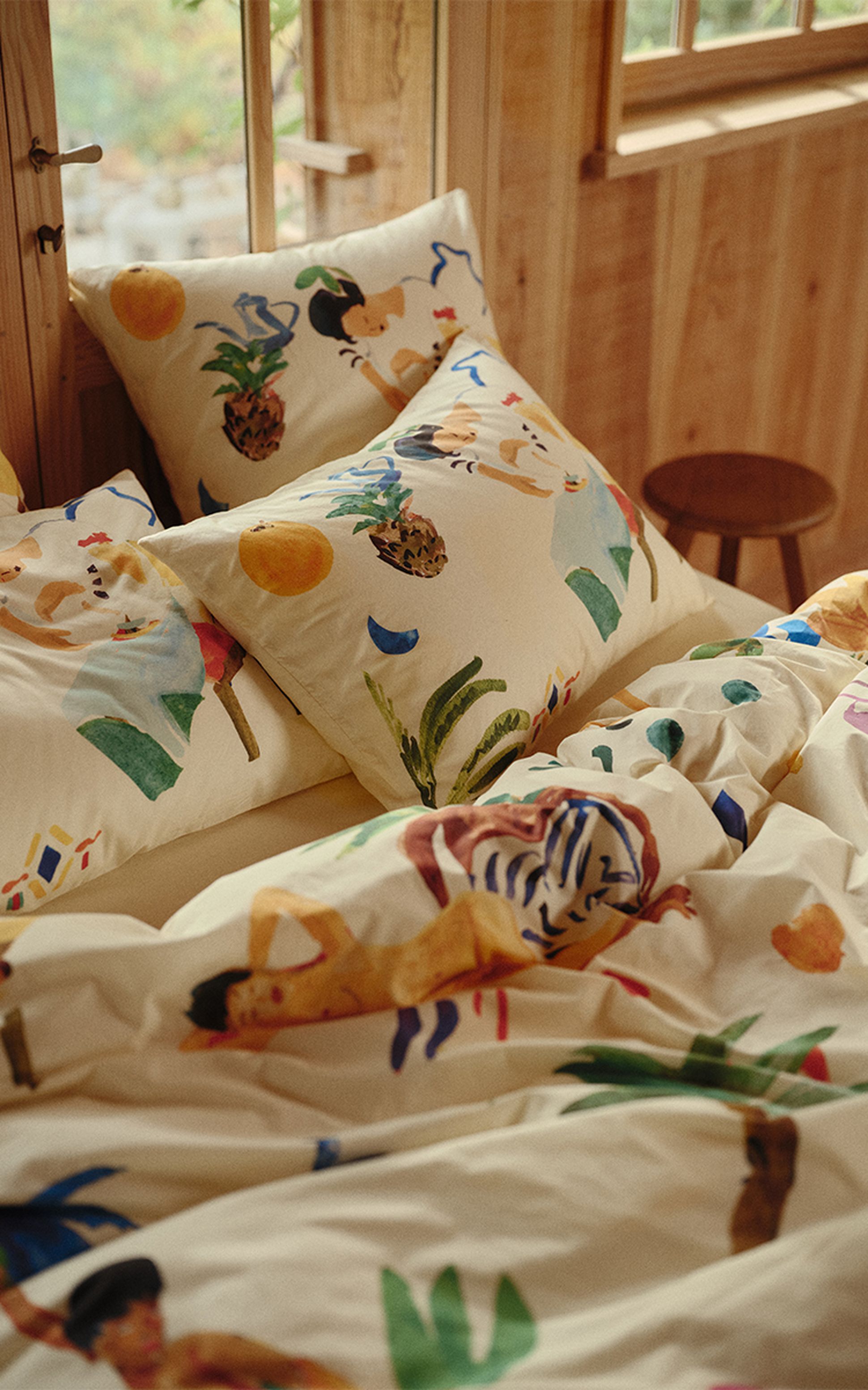 Linge de lit décoratif orné d'illustrations colorées représentant des personnages, des fruits et des plantes, le tout dans une chambre chaleureuse en bois dotée d'une fenêtre et d'un tabouret.